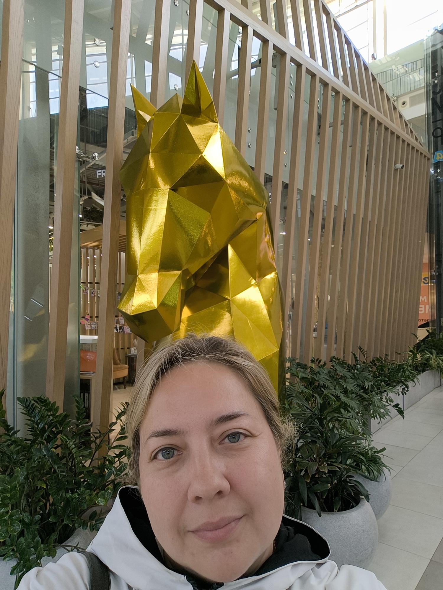 A woman with blonde hair and blue eyes takes a selfie. She is wearing a white hooded jacket with black trim. Behind her, a large, geometric sculpture of a golden horse head is positioned in front of a wooden slatted wall. Plants in grey pots are visible in the foreground and background. The setting appears to be an indoor public space, possibly a shopping mall or atrium, with bright lighting suggesting daytime. The woman has a slight smile and her gaze is directed towards the camera. The overall mood is casual and relaxed. Visible text includes "FR" and a partially obscured sign with "NE" and "M" in orange.