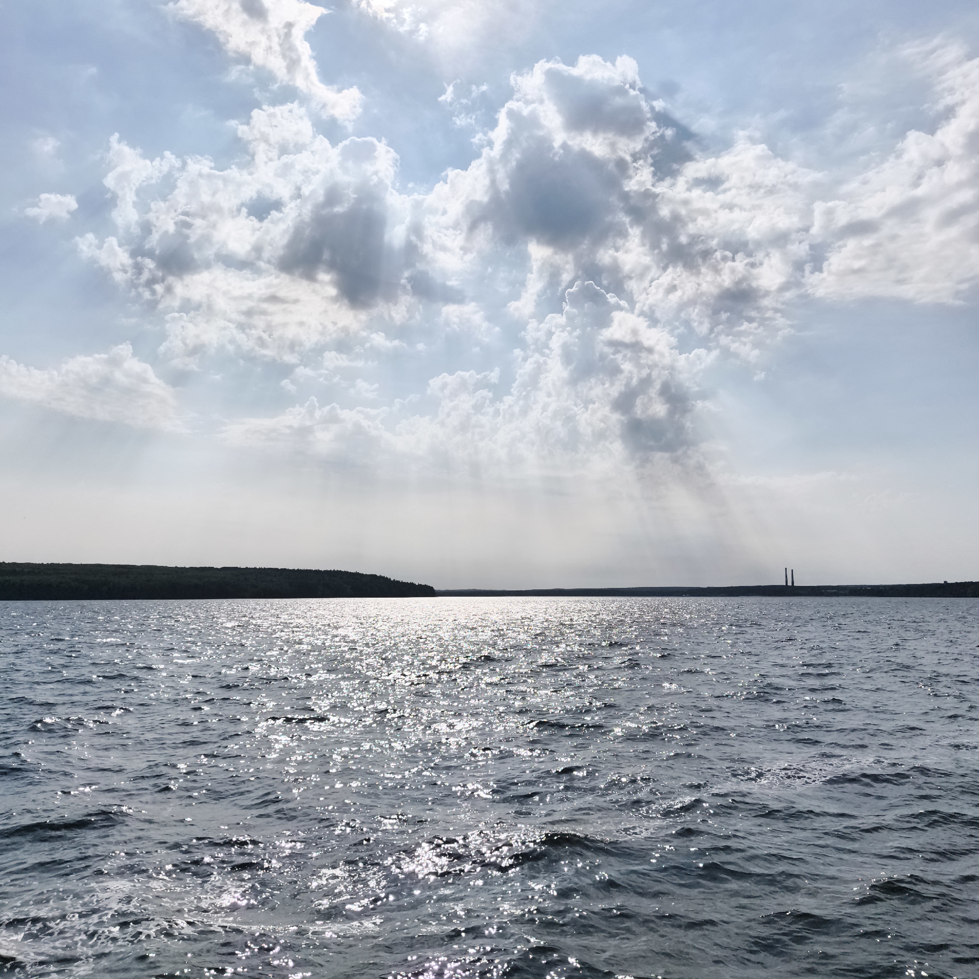 The image depicts a wide body of water, likely a reservoir or a large river, under a partly cloudy sky. Sunlight streams through gaps in the clouds, creating visible rays that illuminate the water's surface, which is choppy and reflects the light in numerous sparkling points. In the distance, a dark, treeline marks the far shore, with two tall, narrow smokestacks visible on the horizon to the right of center. The scene appears to be a natural landscape, with no immediate signs of human activity on the water or the shore, apart from the distant industrial structures. The overall impression is one of a calm, yet dynamic, natural environment. The location is likely near Chaikovsky, Russia, a city known for its Kama River reservoir and associated industrial facilities.