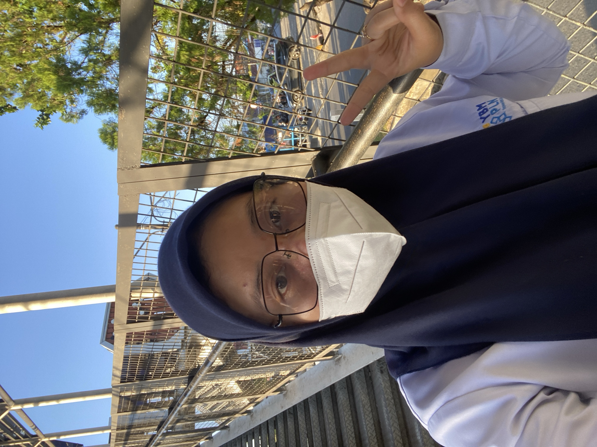 A woman wearing a dark blue hijab, glasses, and a white face mask is taking a selfie on an outdoor pedestrian bridge. She is wearing a light blue long-sleeved shirt with a logo on the left chest that appears to say "YBM PLN." Her right hand is raised, making a peace sign. The bridge has metal stairs on the left and a metal railing with wire mesh on the right. In the background, behind the mesh, is a street with several motorcycles and cars, and some trees and buildings. The sky is clear and blue, suggesting a sunny day. The location is identified as Banjarbaru, Indonesia.