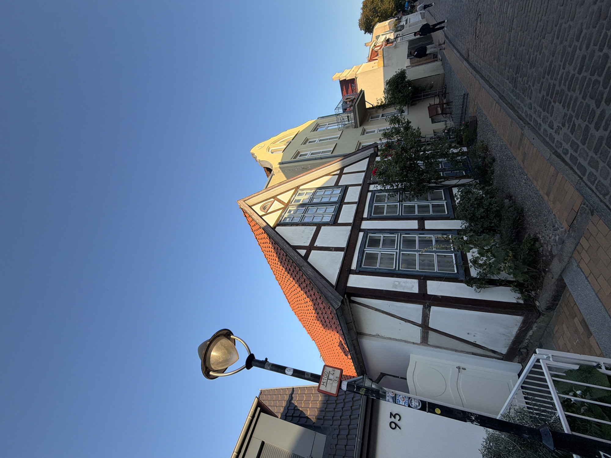 This image captures a narrow cobblestone street lined with historic buildings in Rostock, Germany, likely in the historic city center given the architectural style. The buildings feature traditional German Fachwerk architecture with visible timber framing on a white stucco facade, and red-tiled gabled roofs. One building prominently displays a street number "93" next to a lamppost. The scene is bathed in the warm light of what appears to be late afternoon or early morning sun, casting long shadows across the street and highlighting the textures of the cobblestones and building facades. A few people are visible walking down the street, adding a sense of scale and activity. The sky is clear and blue.