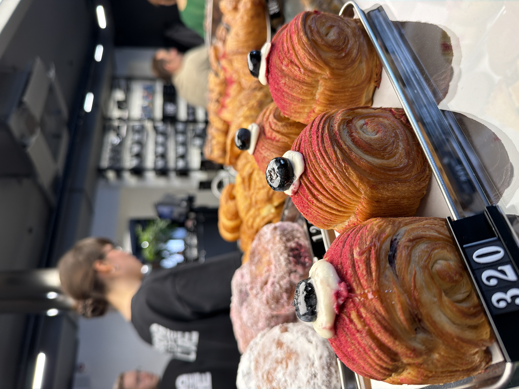 The image displays a close-up, angled view of an assortment of pastries showcased on metal trays within what appears to be a clear display case in a cafe or bakery. The primary focus is on several types of baked goods.
In the foreground, there are multiple spiral-shaped pastries, likely laminated dough creations such as croissants. These have a golden-brown, flaky texture with visible layers. Many are partially dusted or glazed with a vibrant red color, and each is topped with a small dollop of white cream and a single dark berry, resembling a blueberry. Behind these, other pastries are visible, coated in powdered sugar, some appearing plain and others with a reddish or purple hue beneath the sugar, possibly indicating a berry filling. Further back, more golden-brown, spiral-shaped pastries are arranged on additional trays.
The pastries are presented on shiny metal trays, with a clear partition or glass cover partially visible, where a pair of metal tongs rests. A black price tag holder with white numbers is present in the lower right section of the image. The visible text on this tag is "320".
The background is blurred, suggesting a shallow depth of field. It shows elements of a cafe interior: on the left, a person with dark hair styled in a bun, wearing a dark top, is partially visible, looking away from the camera. Other indistinct figures, possibly customers or staff, and various pieces of cafe equipment or shelving units are also discernible. White strip lights are visible on the ceiling.
The overall impression is a bustling indoor commercial setting, likely a cafe or bakery in Izhevsk, Russia, during daytime or evening, with an emphasis on freshly prepared, visually appealing pastries.