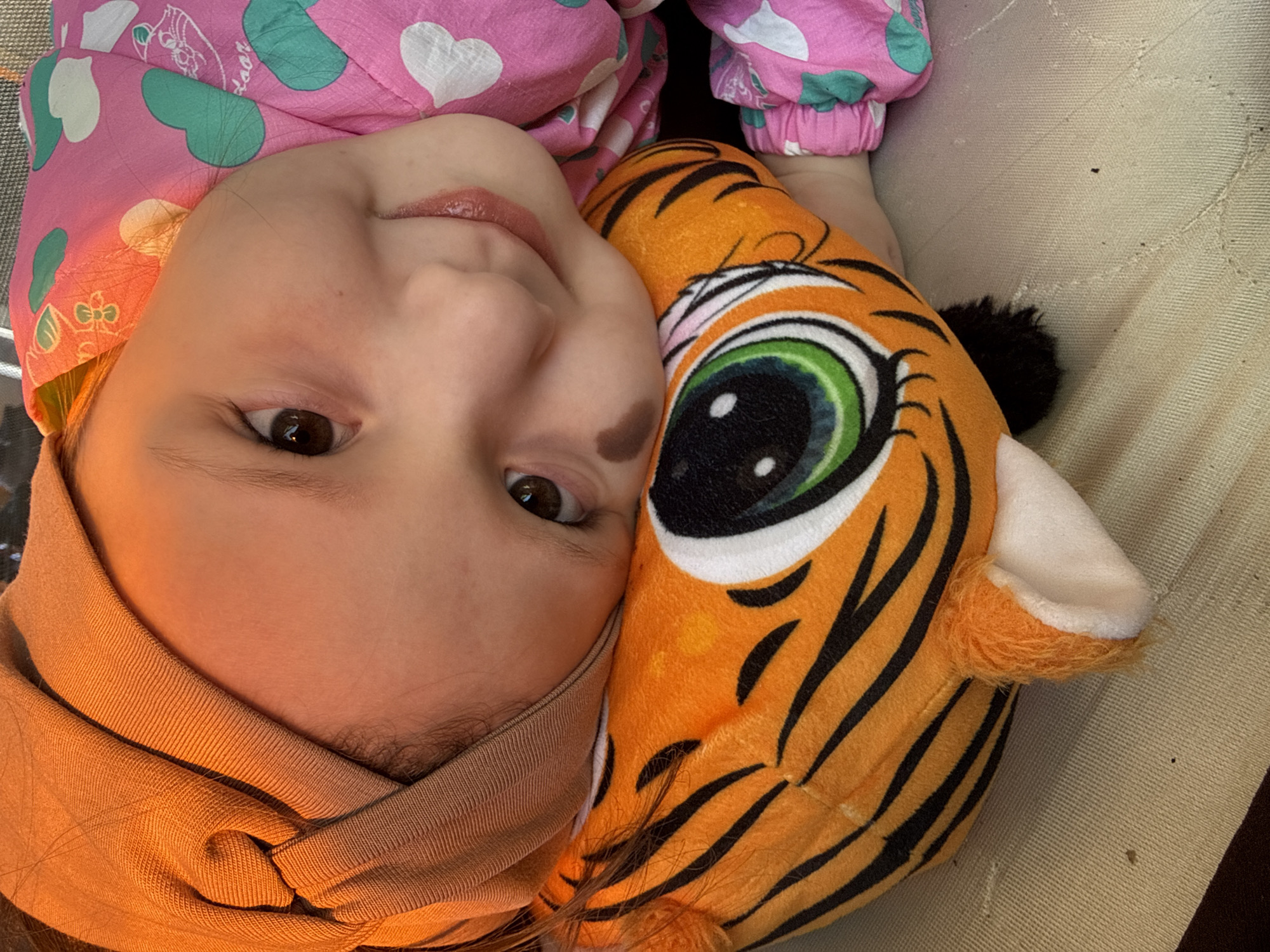 A close-up shot shows a young child lying down with a plush tiger toy. The child has fair skin, brown eyes, and a brown birthmark on her left cheek. She is wearing a light brown headband and a pink jacket with white hearts and green shapes. Her face is partially pressed against the plush tiger, which has orange and black stripes and a large, green and black eye. The background is a beige textured surface, likely a couch or bed. There is no information to discern the location of the scene in the city of Кипчак, Russia, so the location is unknown.