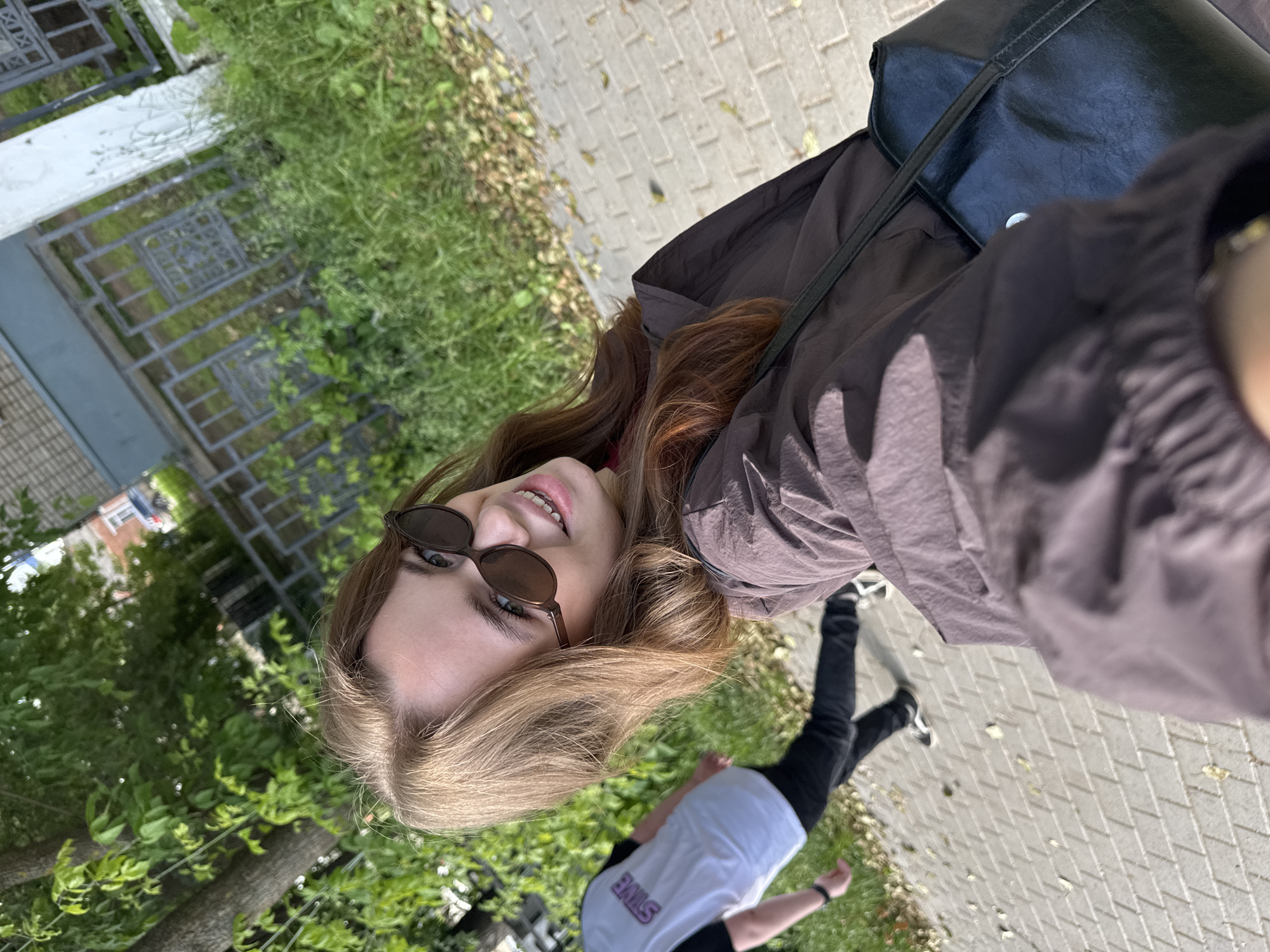 A young woman with long, light brown hair and sunglasses is taking a selfie outdoors. She is wearing a dark brown jacket and a black purse is slung over her shoulder. In the background, another person is walking away, wearing a white t-shirt with "STINE" printed on the back and black pants. The scene appears to be on a paved sidewalk next to a grassy area with trees and a fence. The architecture in the background suggests a residential area, possibly in the city of Chaikovsky, Russia, given the context. The lighting indicates it is daytime, likely sunny.