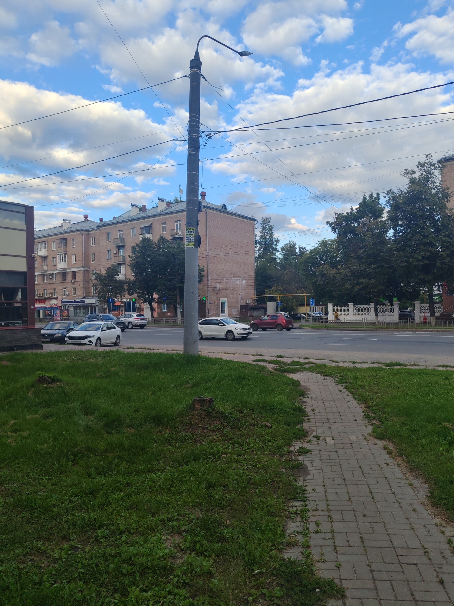 The image depicts a street scene in Nizhny Novgorod, Russia, on a cloudy day. A sidewalk made of grey pavers runs diagonally from the lower right corner into the distance, bordered by a strip of green grass on the left and right. A tall, dark lamppost stands prominently in the foreground, positioned slightly left of center, with electrical wires crisscrossing overhead.
Across the street, a multi-story apartment building with a light brown brick facade dominates the background. The building features multiple windows, balconies, and a grey tiled roof. Several cars are visible on the street: a white sedan is parked near the sidewalk, a dark-colored sedan is parked further back, and a white SUV is parked in front of the building. To the right of the apartment building, a row of trees with lush green foliage lines the street. Further down the street, a white car is driving away from the camera. A street sign with Cyrillic text is attached to the lamppost. The sky is a vibrant blue with scattered white clouds.