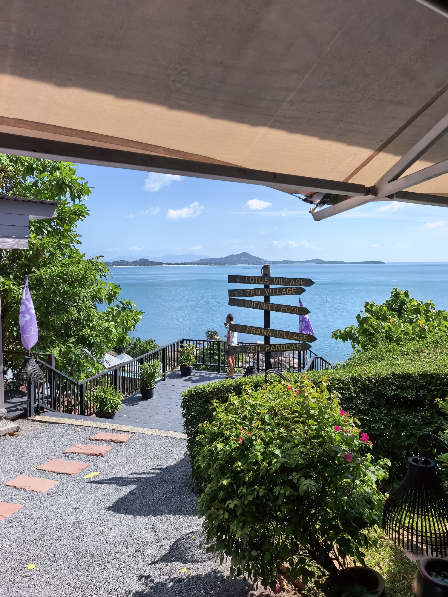 A scenic outdoor view captures a walkway leading towards the ocean on a sunny day. The walkway is paved with gravel and accented with stepping stones, bordered by lush green bushes. A dark railing with an accompanying staircase guides the path downwards. The serene blue ocean stretches to the horizon, dotted with distant islands and a clear sky with scattered clouds.
A signpost with multiple directional arrows stands prominently, indicating various locations: "LOTUS VILLAGE," "ZEN VILLAGE," "INFINITY POOL," "PRANA VILLAGE," and "ZEN PAGODAS." A woman is observed standing near the signpost, facing the ocean, appearing to be taking in the view.
The scene is framed by an awning overhead, providing shade, and the foreground features dense green foliage, some with vibrant pink flowers, and decorative dark metal lanterns. The architecture visible on the left includes a section of a building with grey siding and a purple flag. The overall impression is one of tranquility and a resort-like atmosphere, likely situated in the Ko Samui District of Thailand, given the presence of the signpost's village names.