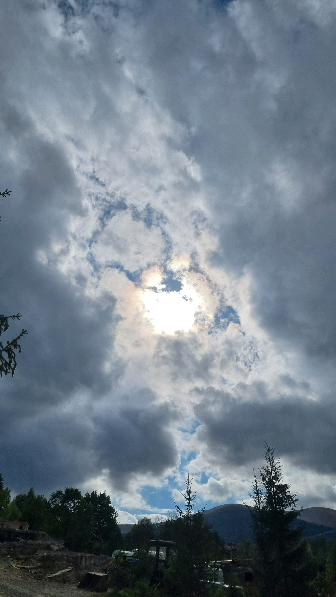 A wide-angle, low-angle, vertical shot captures a dramatic sky over Izky, Ukraine. Dark, heavy clouds dominate the upper portion of the frame, with a bright, sunlit break in the center creating a halo-like effect around the sun, with hints of rainbow colors. The lower third of the image shows a rural landscape with a dark forest and rolling hills in the distance under a partly cloudy sky. In the foreground, there is evidence of construction or land clearing, with debris, machinery, and scattered trees. A large evergreen tree stands prominently on the right, with a smaller evergreen to its left. The lighting suggests it is daytime, possibly late afternoon.