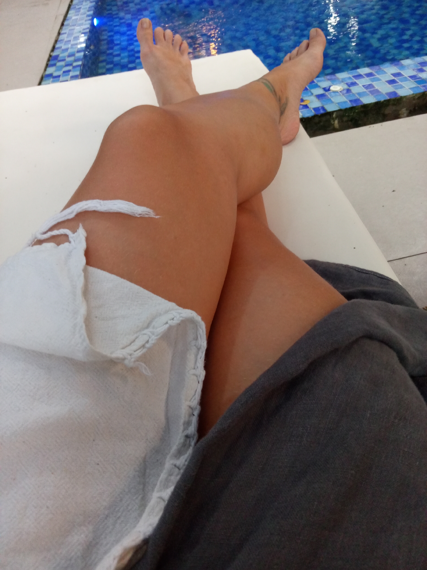 A close-up shot shows a person relaxing on a white lounge chair beside a pool. The person has their legs crossed, with a white garment draped over the left leg and a grey one on the right. The feet are visible at the top of the frame, positioned near the edge of the pool. The pool's tiles are blue and white. The scene is likely in Bac Natural Heritage Area Trang, Vietnam.