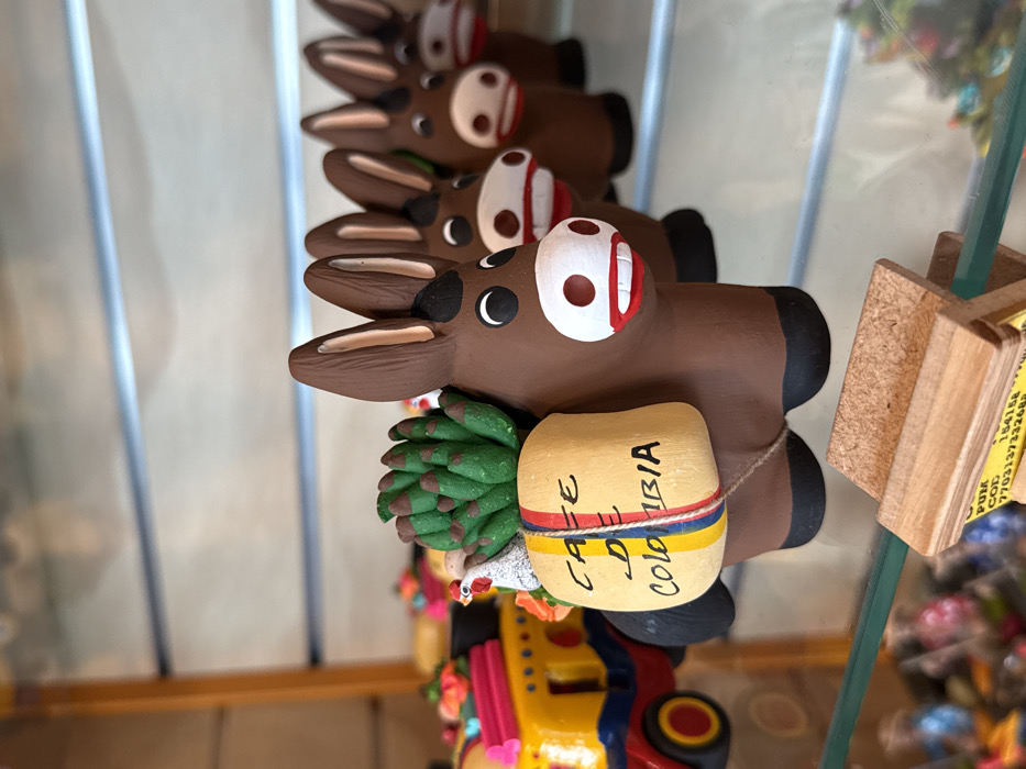 The image displays a clear glass shelf within a retail environment, likely a souvenir shop in the AMCO (Area Metropolitana Centro Occidente) region of Colombia. A primary focus is on a brown ceramic donkey figurine positioned centrally, featuring a white snout with red lips, black eyes, and pointed brown ears with white interior accents. This donkey carries a light beige sack on its back, labeled "CAFE DE COLOMBIA" in black and red text, secured by a string with red, yellow, and blue stripes. A green, spiky plant-like object and a small white chicken figure are placed atop the sack. Behind this lead donkey, a linear formation of identical or similar brown ceramic donkey figurines extends into the background, progressively out of focus, each exhibiting similar facial features. To the left, a portion of a yellow ceramic bus or truck with red wheels and blue details is visible. Below the shelf, a wooden display stand holds a white label partially displaying "PUM COD 154158" and "770313773324". The background comprises additional blurred items on shelves, indicative of merchandise.
