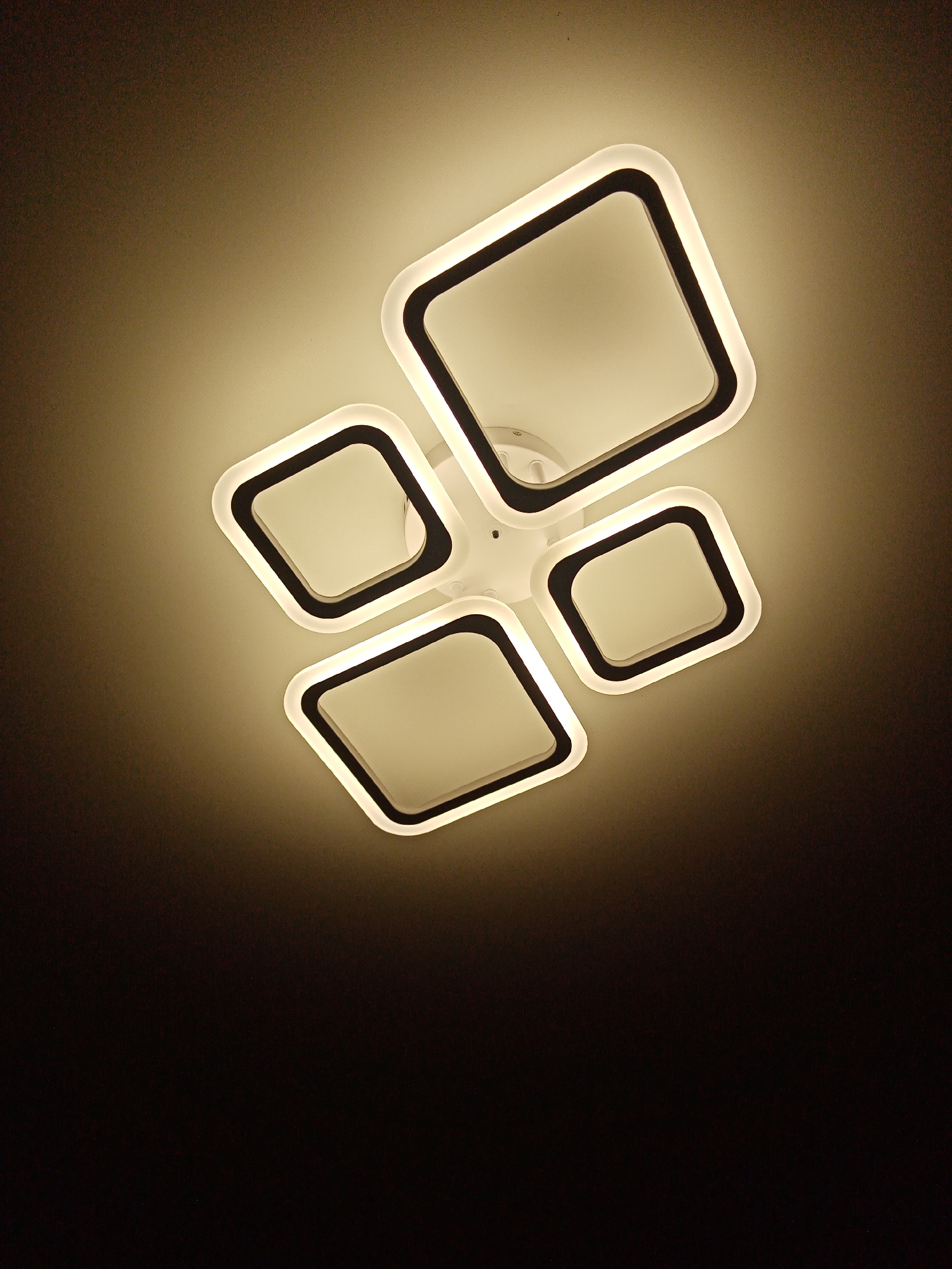 A ceiling-mounted light fixture dominates the upper portion of the image against a gradient background transitioning from a lighter hue at the top to deep shadow at the bottom. The fixture consists of four rounded square shapes arranged asymmetrically. Each square features a black outer border and a white inner section that emits light. The overall color temperature of the light is warm. The central mounting point is a small, round, white element. The perspective indicates the image was taken looking directly upwards at the light fixture. The city of Elektrostal, Russia, is not discernible from the image.