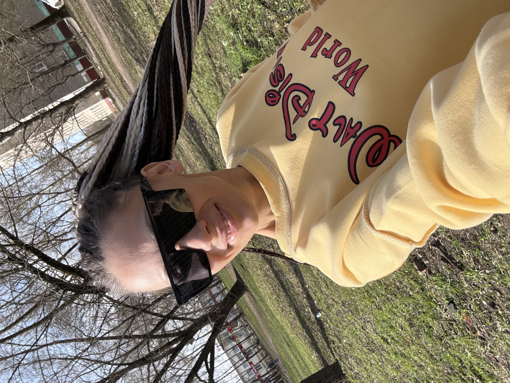 A woman is outdoors, wearing sunglasses and a yellow sweatshirt with "Walt Disney World" printed on it. She has long braids. The background shows a park-like area with trees, grass, and a building in the distance. This image was taken in Fedorino, Russia.