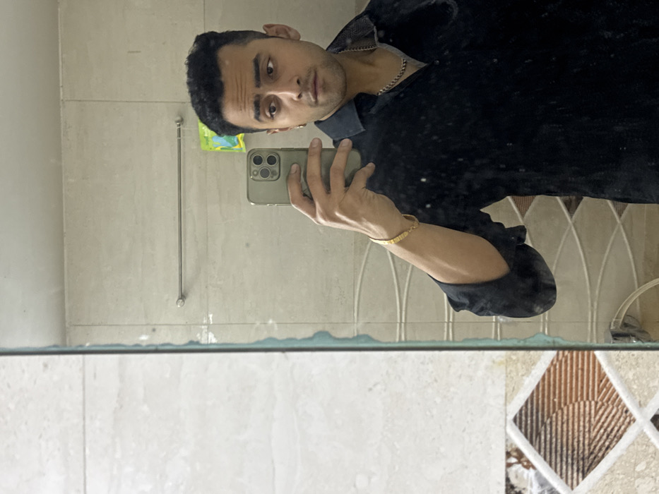 A man in a dark shirt and silver chain takes a selfie in a bathroom mirror. He holds a light green phone, displaying a gold bracelet on his left wrist. The bathroom features light-colored tile walls with a towel rack. The lower portion of the mirror shows a countertop with decorative tile accents. The image is taken at an angle, creating a rotated perspective.