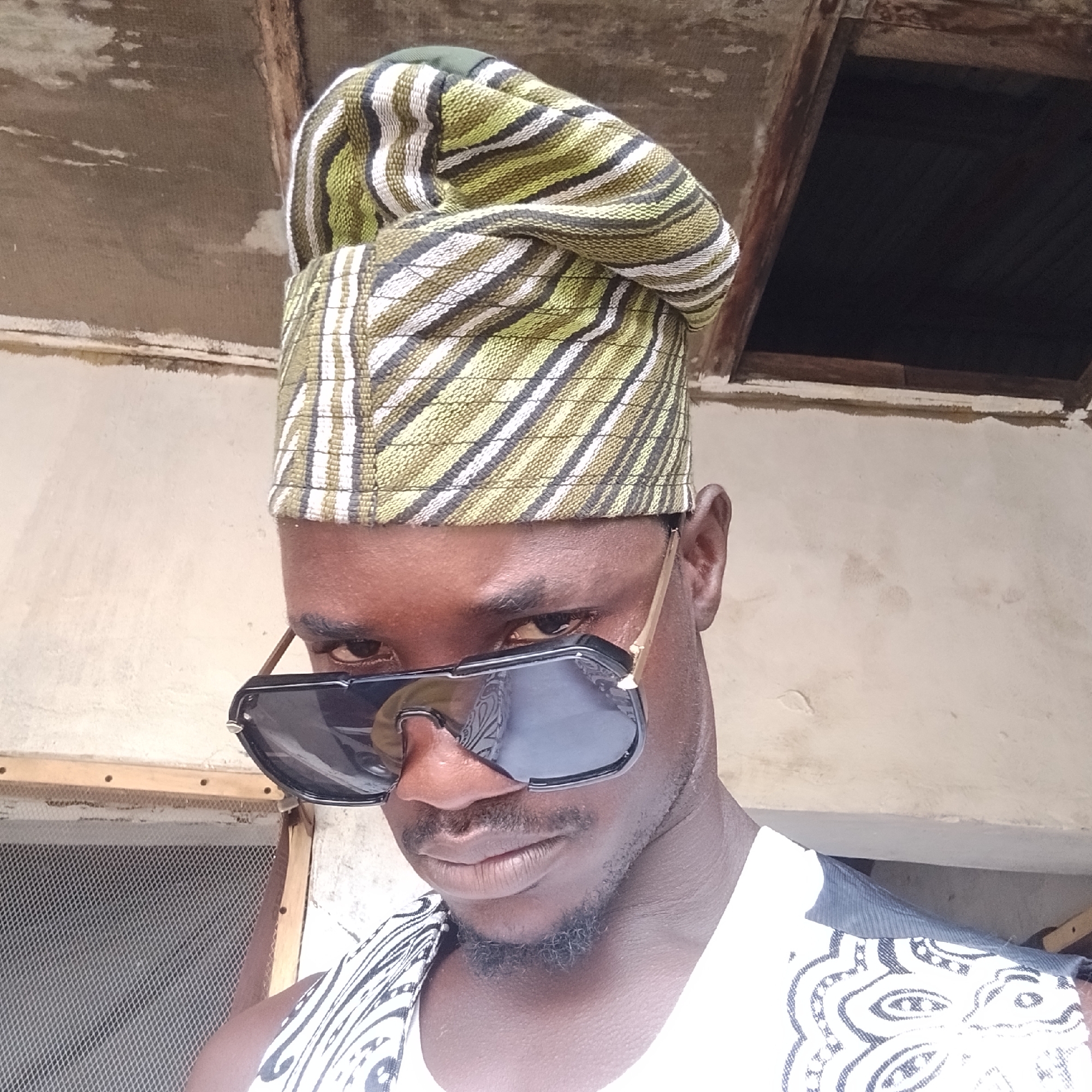 The image shows a close-up, head-and-shoulders shot of a Black male wearing sunglasses and a patterned cap. He is looking down and to his left, with his right eye visible. He is wearing a white garment with a black and white pattern on the shoulder. The background appears to be an outdoor setting with a textured, light-colored wall and a wooden structure overhead. The overall impression is that of a casual portrait taken in an urban environment. Based on the visual information provided, the location cannot be identified as Ado, Nigeria.