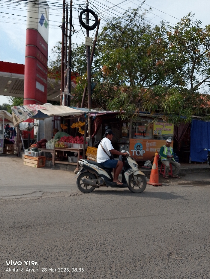 The scene is taken in Jatibarang, Indonesia (latitude: -6.466, longitude: 108.3). A man wearing a white shirt and blue jeans is driving a white motorcycle on the paved road. The motorcycle is heading towards the right. In the background, there is a small shop with a red awning and a sign that says "Bubur Ayam & Soto Ayam". A man wearing a green vest and blue jeans is sitting on a small chair in front of the shop. There are a few crates of fruits and vegetables on the ground near the shop. There is a tall red structure with a blue diamond on it, and a sign that says "SLiM" on the top. The surrounding environment is a small town with trees, power lines, and a few buildings. The sky is clear and blue.