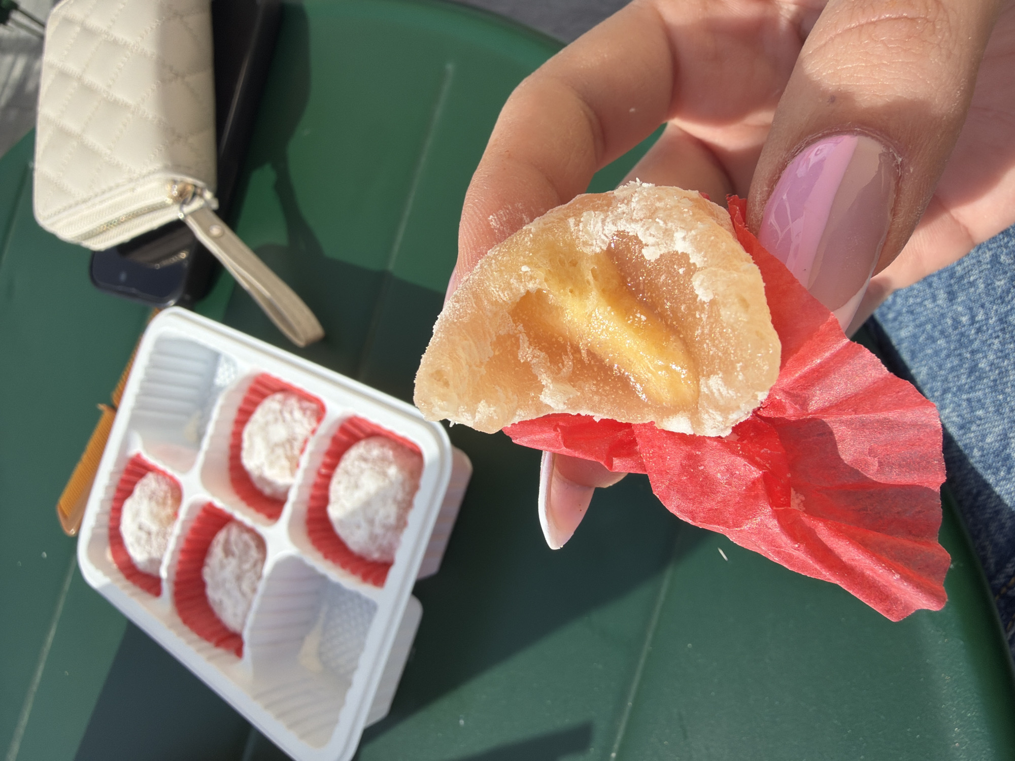A close-up, high-angle shot captures a person's hand holding a sweet outdoors in Veliko Tarnovo, Bulgaria. The central focus is a light-skinned hand, exhibiting a two-tone pink and light beige French manicure on the fingernails, holding a piece of translucent, sugared candied fruit or mochi with a lighter yellow interior, partially wrapped in a crinkled red paper liner. The sweet itself shows a cross-shaped indentation on its surface.
In the upper left quadrant, a white plastic tray with six rectangular compartments holds five small, round sweets, each dusted with a white powder and placed in individual red paper liners. One compartment is empty. To the upper right, a cream-colored, quilted zipper wallet with a wrist strap rests atop a dark-colored smartphone. All objects are positioned on a dark green, slightly textured surface, indicative of a table or bench, under direct sunlight, which casts distinct shadows. The lower portion of the image reveals blue denim fabric, suggesting the person is wearing jeans. The specific street or landmark in Veliko Tarnovo is not visually discernible.