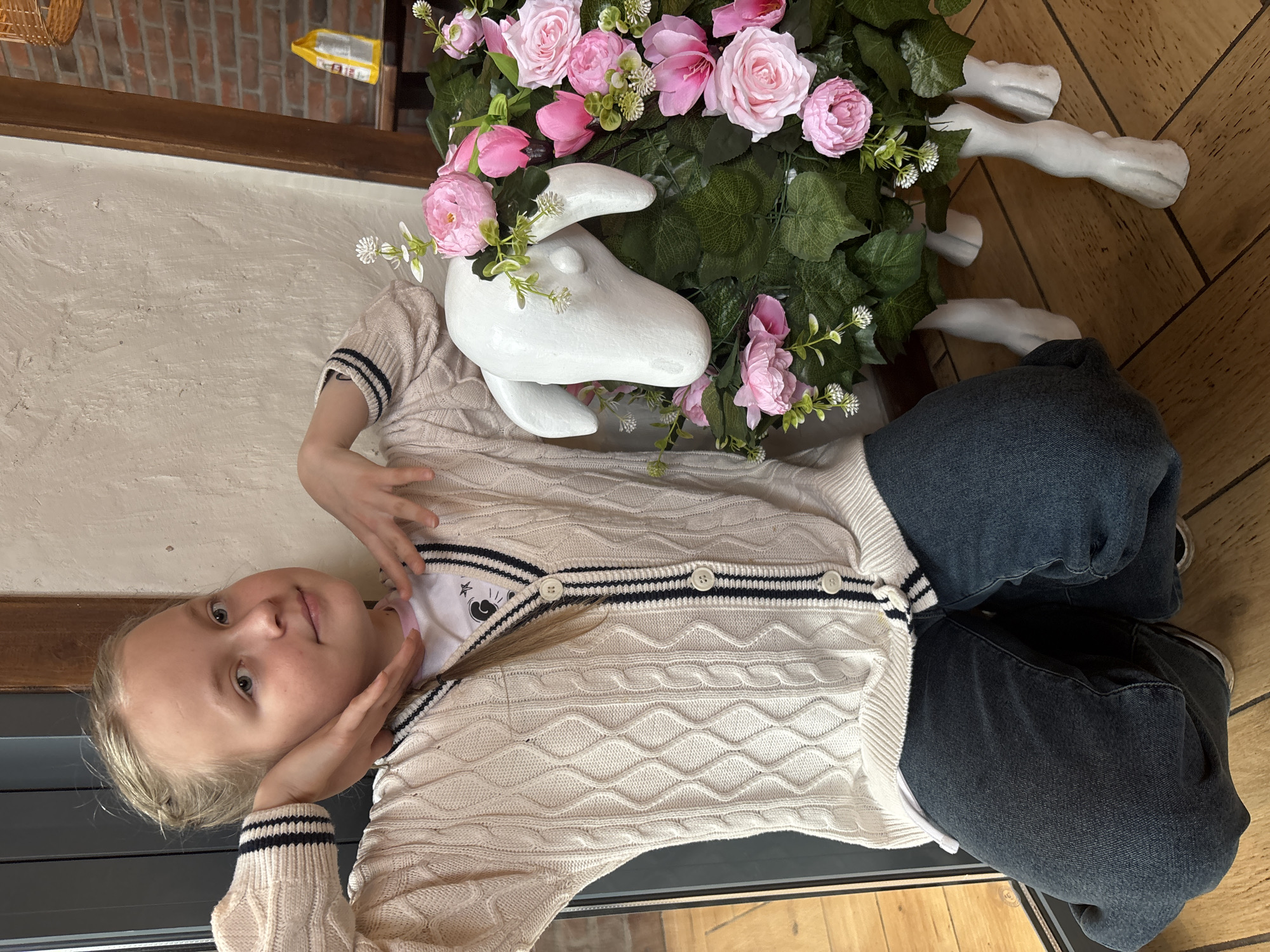 A blonde-haired child lies next to a white, sheep-shaped decorative object adorned with pink roses and greenery. The child wears a white cardigan with black stripes, a t-shirt with a cartoon graphic, and dark denim jeans. They are lying on a wooden floor with their hands near their face. The background features a brick wall and what appears to be a hanging object. There is no information to indicate that this photo was taken in Izhevsk, Russia.