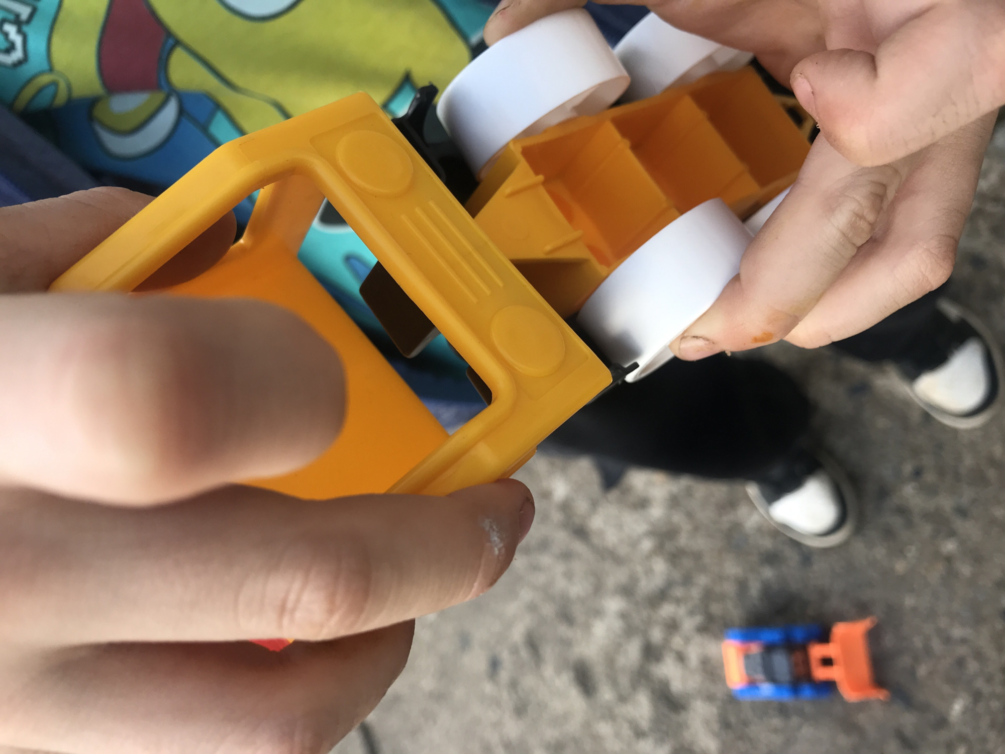 The image presents a close-up, high-angle view of hands interacting with a brightly colored plastic toy, set outdoors in Chaikovsky, Russia. In the foreground, a person's hands, likely those of a child, hold a yellow plastic toy. This toy features a sturdy yellow frame with circular raised details and linear grooves on its surface.
Further into the frame, another set of hands is also engaged with the toy. The toy itself appears to be a construction-type vehicle, comprising the prominent yellow plastic structure, white cylindrical components (resembling wheels or drums), and orange cuboid sections that suggest a cargo bed or container. The hands are fair-skinned, with some smudges of dirt visible, consistent with outdoor play.
In the blurred background, a person's feet, clad in black pants and black and white shoes, stand on a textured grey ground surface, which appears to be paved or gravel. Nearby, a smaller, out-of-focus toy truck, distinguished by its blue body and orange scoop, rests on the ground. Behind the hands and toy, a colorful, patterned fabric, possibly clothing or a blanket, with blue, yellow, and white shapes, adds a vibrant element to the background.
The scene depicts children engaged in outdoor play with toys, likely in a casual, daytime setting given the bright illumination. The focus is on the tactile interaction with the toys. No visible text is present in the image.