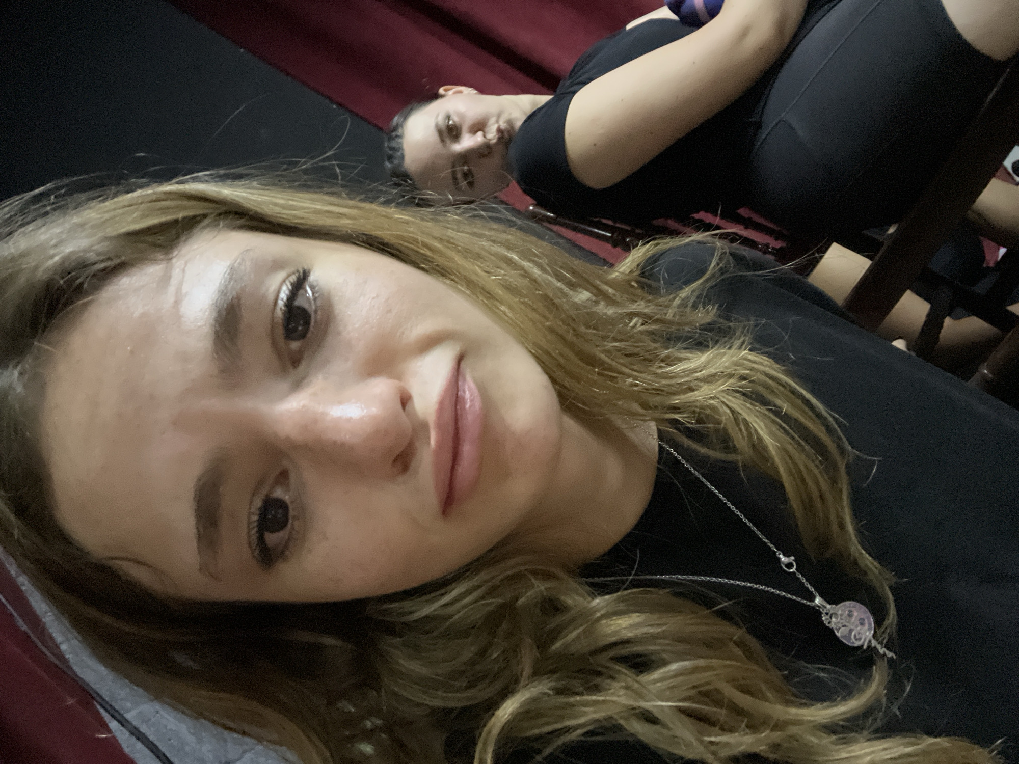 Two females are in the shot, one is in the foreground while the other is in the background, in what appears to be an indoor setting. The woman in the foreground has light brown hair and a silver necklace with a light-colored pendant. She is wearing a black shirt. The other woman is behind her, wearing a black shirt and black athletic pants and seated in a black chair. The setting behind them appears to have a maroon curtain. The location is specified as San Bernardino Tlaxcalancingo, Mexico.