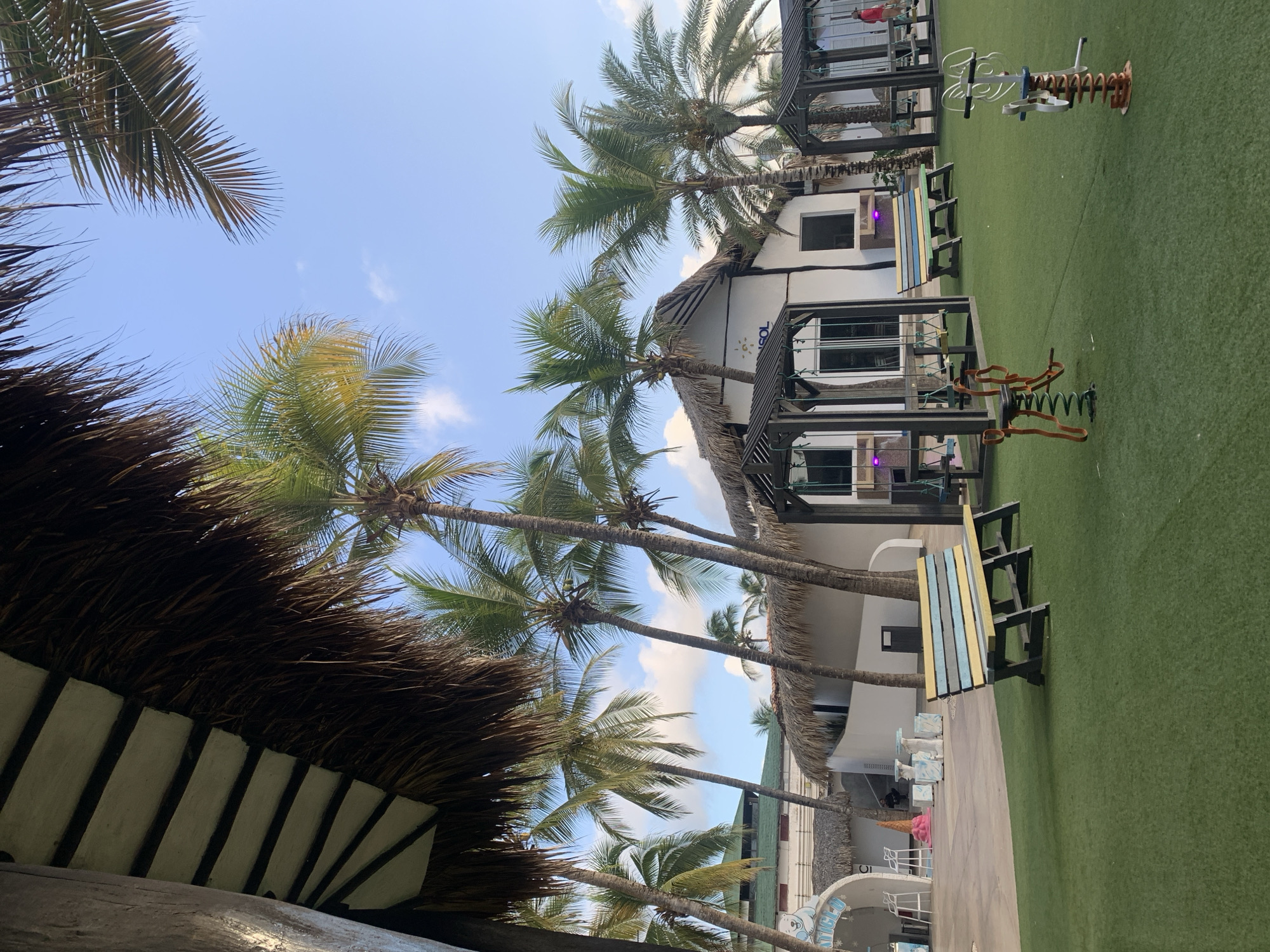 The image shows an outdoor area with artificial grass and palm trees, suggesting a tropical or resort-like setting. In the foreground, there are several pieces of playground equipment, including a red spring rider and a metal see-saw-like toy. A yellow, blue, and green striped bench sits on the grass. Behind the bench, there are covered picnic tables with seating.
In the mid-ground, a white building with a thatched roof is visible, featuring large windows and an arched entranceway. The building appears to be part of a commercial establishment, with a sign that has a sun logo and the text "SOL." Further left, another white structure with a thatched roof has an arched entrance with the word "ATTICLE-U" visible above it, and what appears to be a counter or service area inside.
The background is dominated by numerous palm trees, some with green fronds and others with drier, brown fronds. The sky is blue with scattered white clouds. The overall impression is of a relaxed, recreational outdoor space, possibly a beach resort or a park in El Tirano, Venezuela, given the presence of palm trees and the general climate implied by the scene. There are no discernible people actively engaged in activities, though a single person in a red shirt is partially visible to the far right. The lighting suggests it is daytime, possibly late afternoon due to the angle of the shadows.