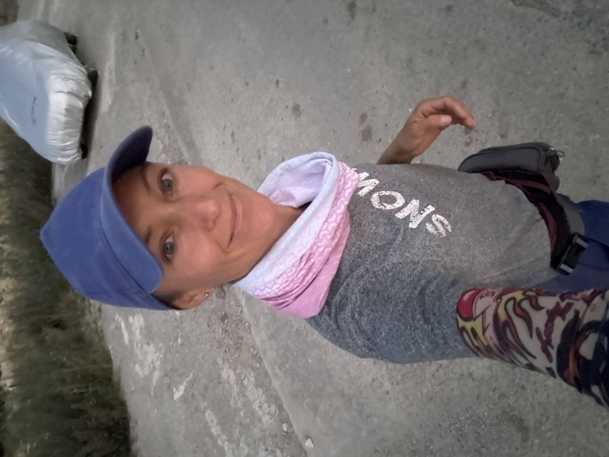 A woman is taking a selfie outdoors. She is wearing a blue baseball cap, a grey t-shirt with white text that reads "SNOW," and a pink and white patterned neck gaiter. She also has a small dark pouch attached to her waist. Her left arm is visible in the foreground, and it appears to be covered by a sleeve with a colorful, flame-like pattern. In the background, to the right of the woman, a car covered in a grey tarp is parked. The ground is paved with concrete, and there is vegetation in the background. The lighting suggests it is either early morning or late evening. This scene is located in Malyi Mayak, Ukraine.
