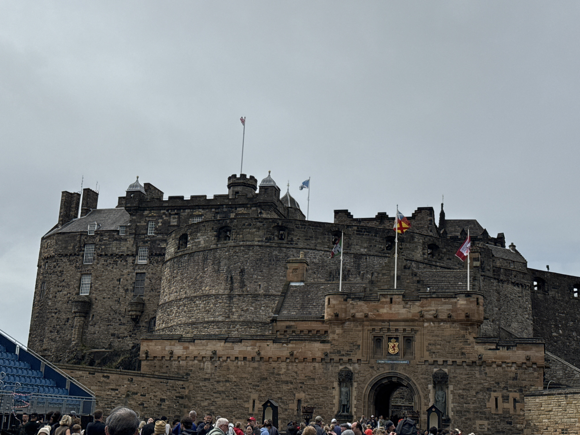 The image shows the exterior of Edinburgh Castle, a historic fortress located on Castle Rock in Edinburgh, Scotland, United Kingdom. The castle is a prominent landmark and a popular tourist attraction. In the foreground, a crowd of people is gathered, appearing to be waiting or observing. Some people are wearing casual clothing, suggesting a daytime visit. The castle itself is a massive stone structure with multiple towers, battlements, and windows. Several flags are visible flying from the castle's ramparts, including what appears to be the Union Jack and the Scottish flag. The sky is overcast, creating a somewhat muted light over the scene. To the left of the image, a section of blue stadium seating is visible, possibly indicating a performance or event being held at or near the castle. The overall impression is of a historical site bustling with visitors on a cloudy day.