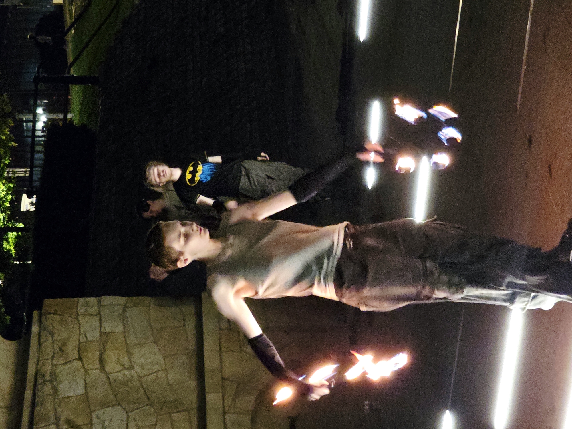 The image depicts a nighttime outdoor scene, possibly in a park or public space in Warsaw, Poland, given the architectural style of the stone wall in the background which resembles some older structures in the city.
In the foreground, a person is engaged in fire performance, holding at least three flaming props in their hands, creating bright streaks of light and flame that illuminate the immediate area. The performer is wearing a sleeveless top and dark pants, with black arm coverings. Their body is angled, suggesting movement and dynamic action.
Behind the main performer, two other individuals are visible. One, to the left and slightly behind, is partially obscured and appears to be looking towards the performer. Another individual, to the right, is wearing a black t-shirt with a yellow Batman logo and blue accents. They are also looking in the general direction of the performance.
The environment is dark, with the primary illumination coming from the performer's fire props and some linear light sources on the ground, possibly part of an installation or pathway lighting. The background is indistinct, with shadows and some muted green foliage, hinting at trees and vegetation. The overall atmosphere is one of a public performance or event taking place after dark.