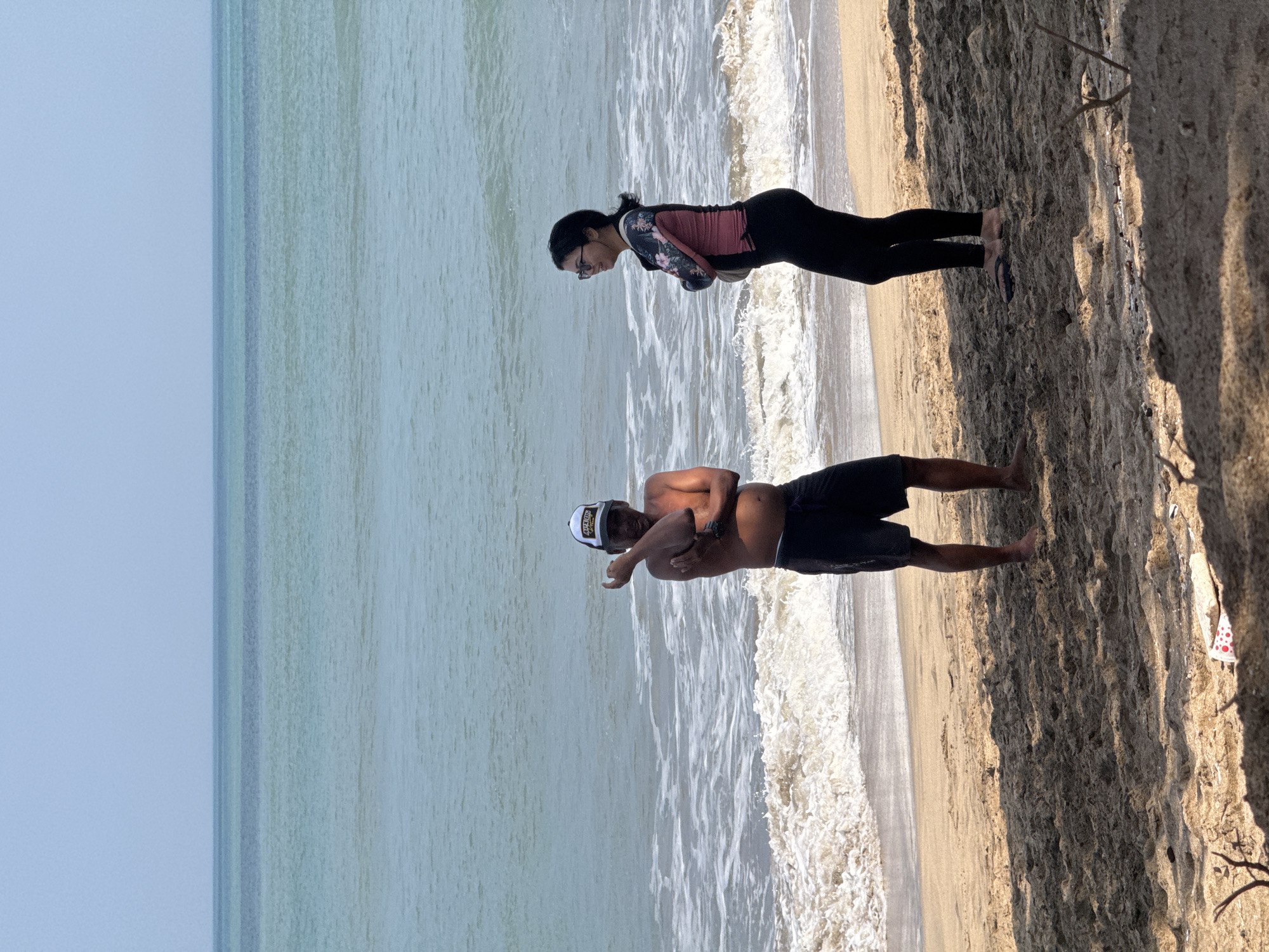 Two people are standing on a sandy beach in Kuta, Indonesia, near the ocean. The person on the left is a male wearing black shorts and a white and yellow baseball cap, with his arms crossed in front of him. The person on the right is a female wearing a pink and black long-sleeved swimsuit and black leggings, with her arms crossed. The ocean is visible in the background with waves breaking on the shore. The sand is light brown and there are some shadows cast on the sand from unseen objects. The sky is a clear light blue.
