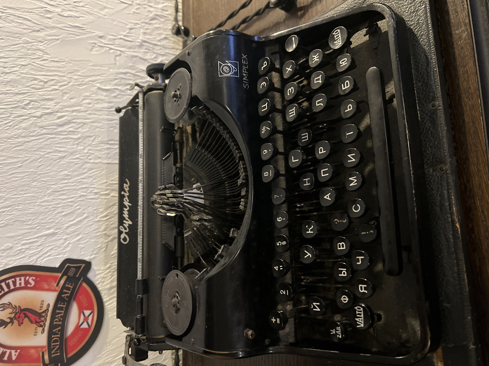 A close-up shot depicts a vintage black typewriter, positioned against a textured white wall and a dark wooden surface. The typewriter, identified as "Olympia" on its left side, also features "SIMPLEX" branding. The keys are Cyrillic, with some labeled "VÁLTÓ". A beer label, "ALEITH'S INDIA PALE ALE" can be seen in the lower left corner. There is no indication of it being from St. Petersburg, Russia, or of any people present.