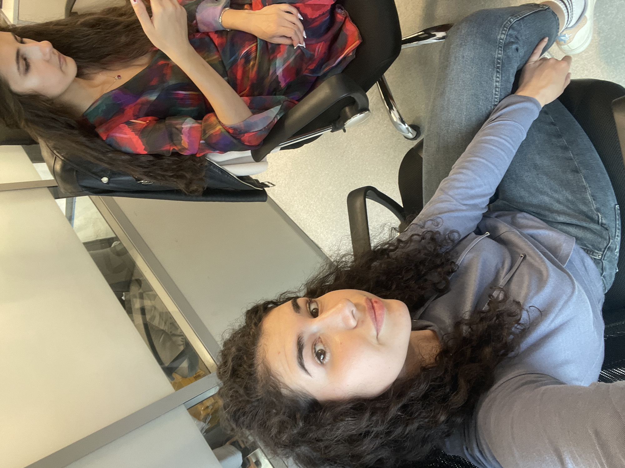 Two young women are captured in an indoor selfie, likely taken in an office or educational setting in Sofia, Bulgaria. The image is rotated 90 degrees to the left.
In the foreground, a woman with voluminous dark curly hair and a light complexion looks directly at the camera with a subtle smile. She is wearing a light blue-grey long-sleeved top and dark blue jeans. Her hands are not fully visible, but she appears to be holding the camera for the selfie.
Behind her, to the upper left of the frame, another woman is partially visible, focusing on her phone. She has long, straight dark hair and a light complexion. She is dressed in a striking long-sleeved top featuring an abstract pattern with vibrant shades of red, purple, green, and black. A delicate necklace with a dark pendant is visible around her neck. Her hands are holding her phone.
Both women are seated in black office chairs with chrome bases. A black bag or backpack is visible on the floor next to the woman in the background. The environment features light-colored walls or partitions, some with reflective or glass elements, indicating a modern interior. The floor is a light, neutral color. The lighting is bright and even, typical of an indoor daytime setting. There is no visible text in the image.