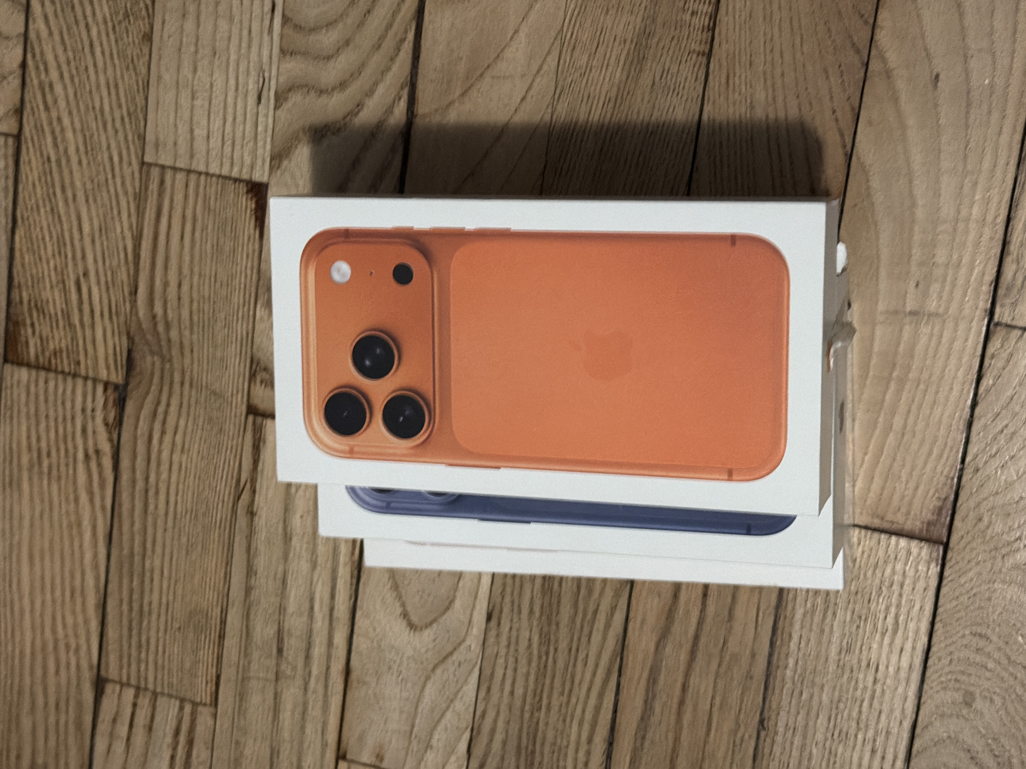 Two iPhone boxes stacked on a wood floor. The top box is orange, and the bottom box is blue.