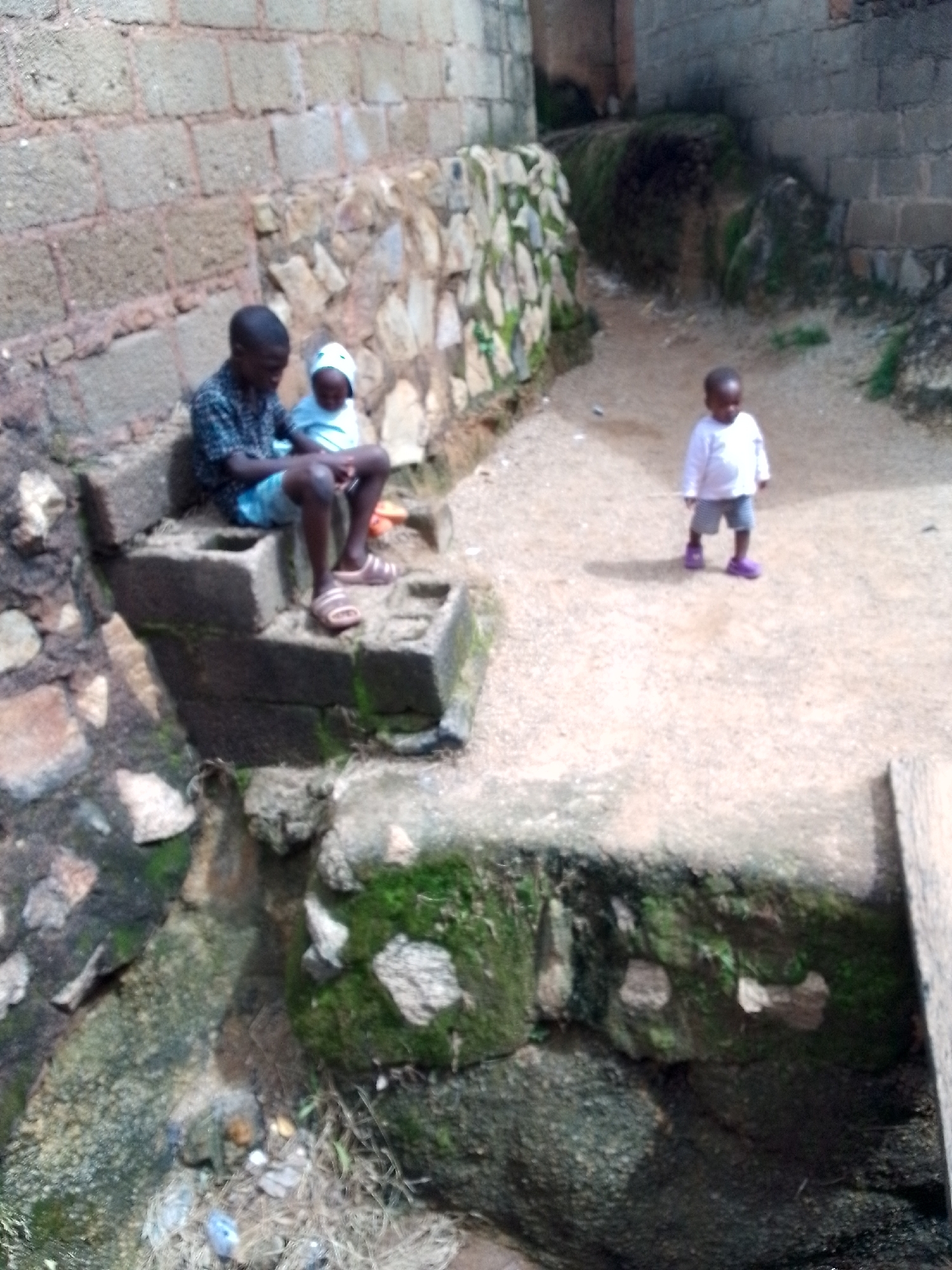 In an outdoor urban environment, two young boys are present. The older boy, dressed in a dark patterned short-sleeved shirt and light blue shorts, sits on a stone structure on the left side of the image. He is wearing tan sandals. Beside him, a younger boy, wearing a white long-sleeved shirt and gray shorts, is also seated. He has a white cap on his head and is wearing light blue sandals. To the right of the seated boys, a much younger child, a toddler, stands on a concrete path. The toddler is wearing a white long-sleeved shirt, gray shorts, and purple shoes. The scene appears to be a pathway or courtyard between buildings constructed with concrete blocks and stone. Green moss grows on some of the stone and concrete surfaces. The ground is a mix of dirt and concrete.