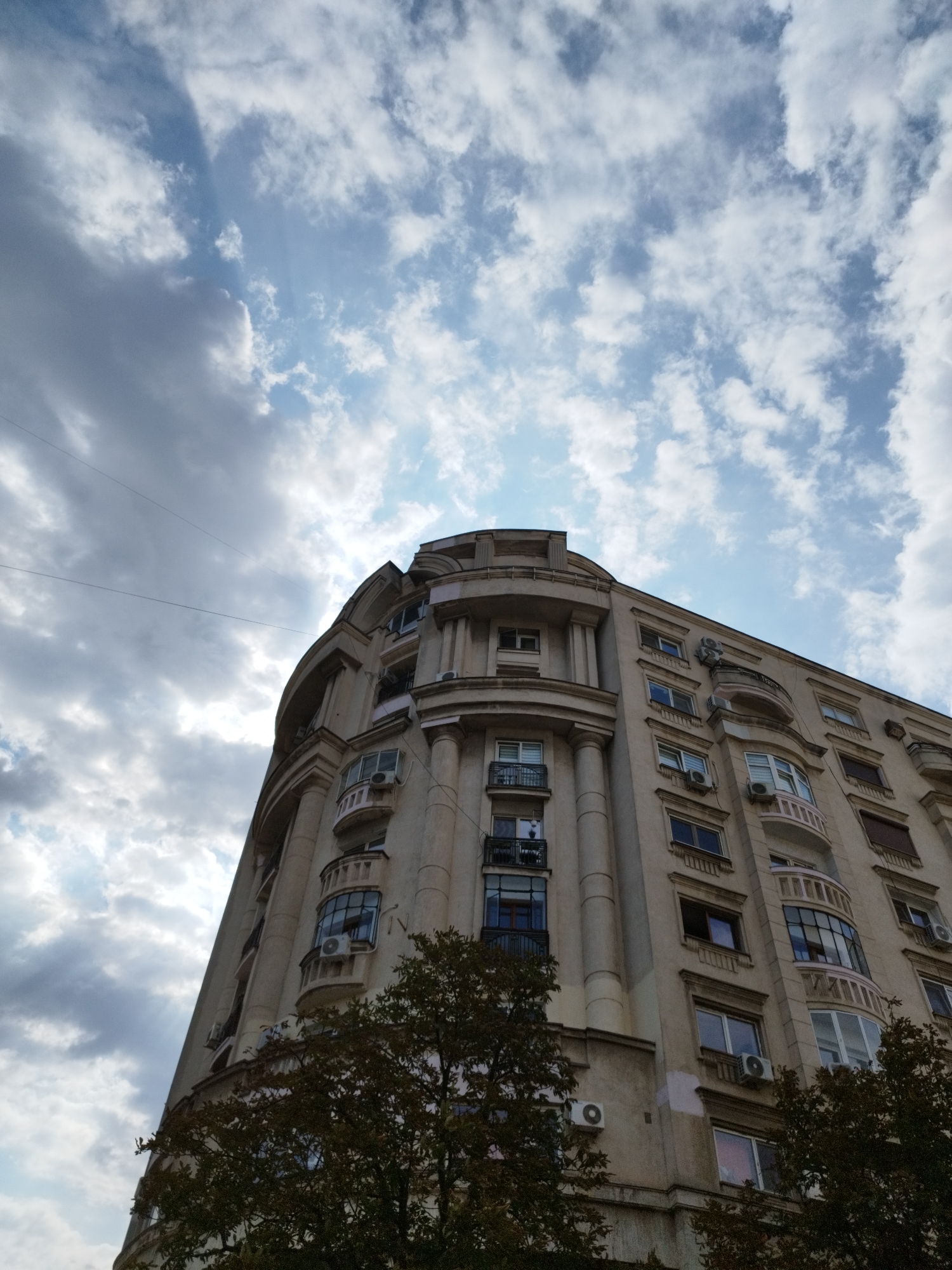 The image is a low-angle, outdoor photograph of a large, beige, multi-story building. The building features a curved facade and numerous windows, some with balconies. Several air conditioning units are visible mounted on the exterior walls. The building appears to be in an urban setting, with trees in the foreground obscuring the lower portions of the structure. The sky above is partly cloudy, with patches of blue sky visible between dramatic gray clouds. The architectural style suggests the building may be located in Bucharest, Romania, possibly in a central or older district known for its apartment buildings. No people are visible in the image. The overall impression is one of a cloudy day in a European city.