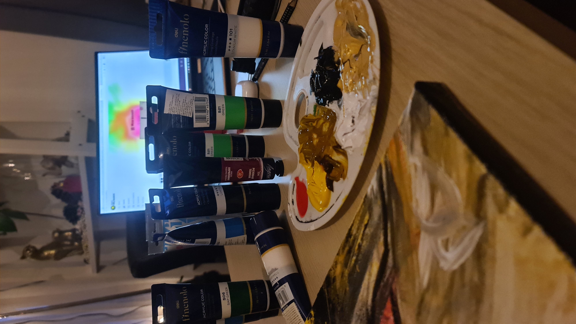 The image shows a tabletop surface, likely made of wood, covered with art supplies. The supplies include numerous tubes of 'deli finenolo' brand acrylic paint in various colors such as titanium white, sap green, dark pink, and green. A white plastic paint palette is also present, holding dollops of black, brown, white, yellow, and red paint. A painting, displaying visible brushstrokes and hues of yellow, white, and dark colors, rests on the table. In the background, out of focus, there is a television screen and some decorative items on shelves. There is no way to tell where this is.