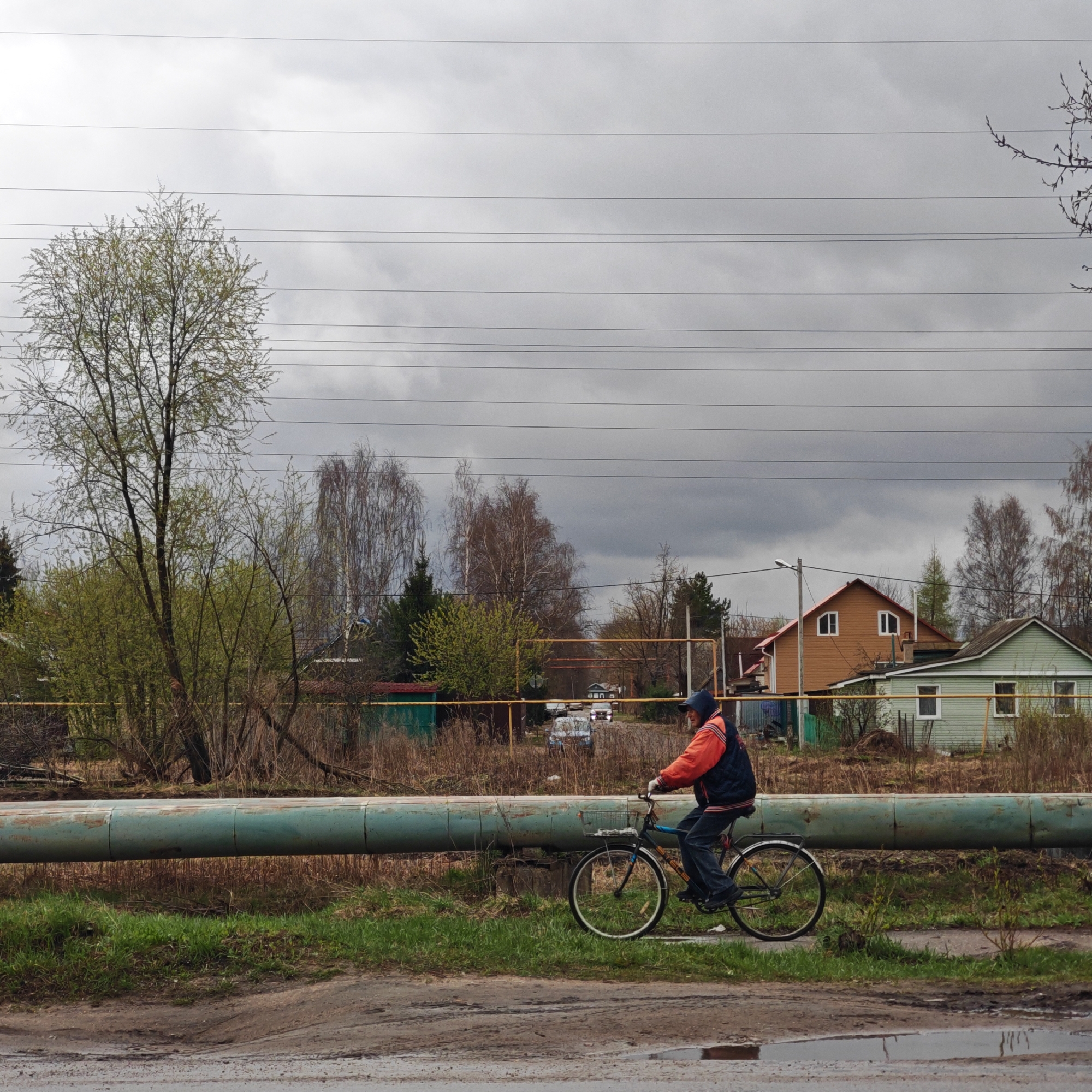 An adult is seen riding a bicycle from left to right across the frame in what appears to be a semi-rural or older residential area in Rostov, Russia. The individual wears a dark blue hooded vest over an orange or red long-sleeved jacket and blue jeans, with a blue cap. The bicycle has a dark frame, white-rimmed wheels, and a rear rack.
A prominent feature in the mid-ground is a large, weathered, teal-colored pipeline running horizontally across the entire width of the image, elevated slightly above the ground. Below the pipeline, the ground is covered with dry, tall grass and patches of green grass. The immediate foreground shows a dirt path or road with puddles and tire tracks, indicating recent rain or a damp environment.
In the background, several houses are visible through the trees. A two-story light brown house with distinct arched white windows stands out, next to a lighter green single-story house. Various trees with budding green leaves and some still bare branches populate the scene, suggesting spring. Yellow metal structures, possibly fences or supports, are scattered in the mid-ground behind the pipeline. Overhead, numerous power lines or telephone wires stretch across a uniformly overcast, grey sky, contributing to a muted and slightly gloomy atmosphere. There is no visible text in the image.