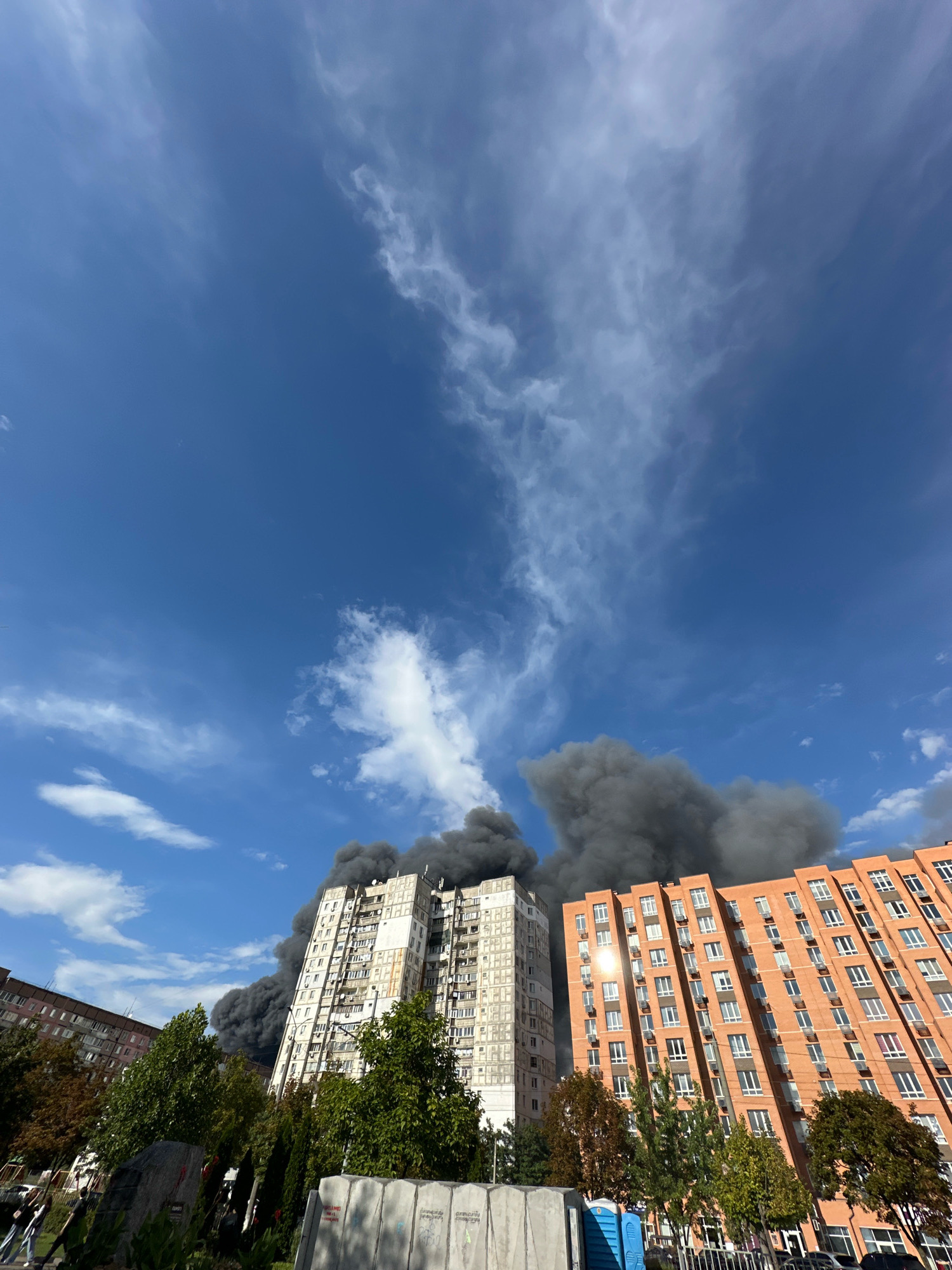 A daytime, wide-angle, low-angle shot captures a residential area in Dnipro, Ukraine, under a bright blue sky with scattered white clouds. The dominant feature is a thick plume of dark grey smoke billowing from the upper floors of a tall, light-colored apartment building located in the center of the frame. To the right of this building is a bright orange, modern apartment building with numerous windows. In front of these structures, a line of trees with green and brown foliage is visible, and further in the foreground, there are some concrete barriers and two blue portable toilets. The scene suggests a fire or a significant incident occurring within the light-colored apartment building, with the smoke obscuring a portion of its facade and drifting towards the orange building.