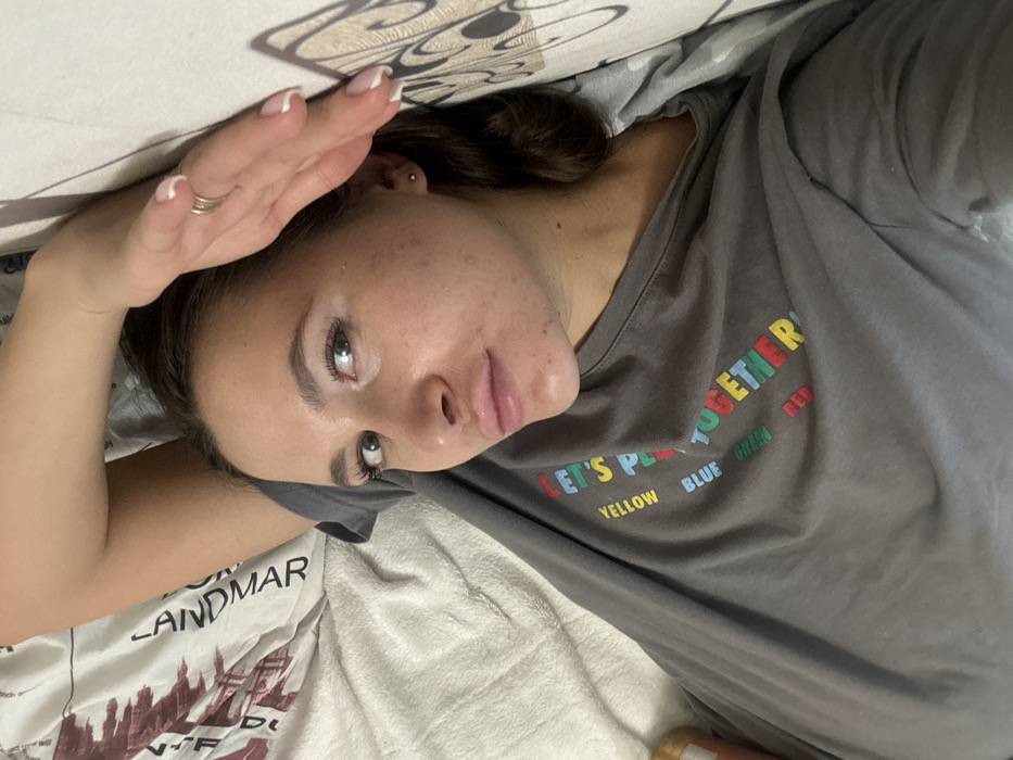 A woman lies on her back, partially obscured by bedding printed with building silhouettes and the word "LANDMAR". She wears a grey t-shirt that reads "LET'S PLAY TOGETHER" with each word in a different color. Her hand is raised above her face, shielding her eyes. She has fair skin with freckles and wears a ring on her left ring finger. The image is taken from a slightly above and angled perspective.