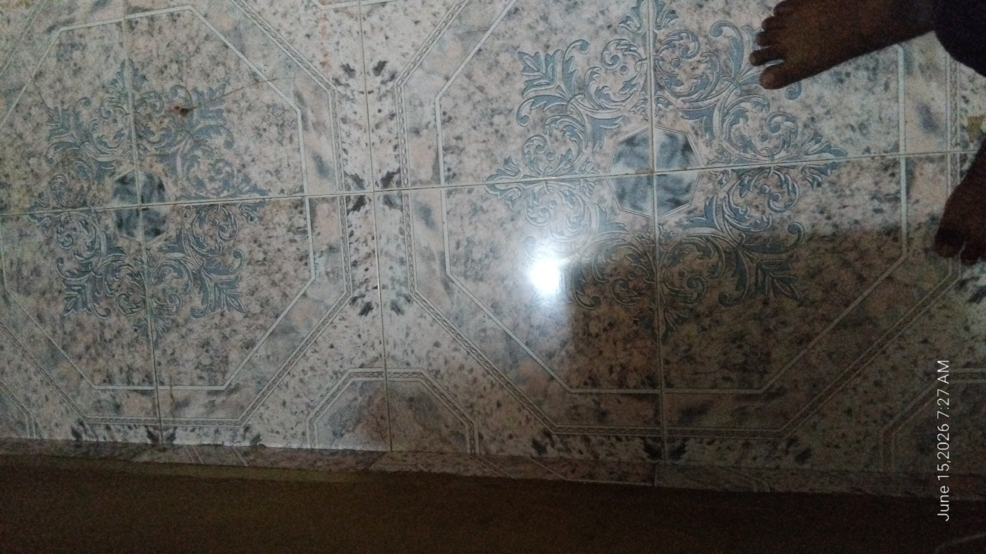 A close-up, overhead view of patterned floor tiles in a domestic setting. The tiles are octagonal and square with a mottled grey and beige base color. An ornate, blue-grey floral or scroll pattern is printed on some of the tiles. The lighting is dim, with a bright glare reflecting off the tiles in the center of the frame. A person's feet are visible in the lower right corner of the image. The timestamp "June 15, 2026 7:27 AM" is overlaid on the bottom left corner of the image. The environment appears to be a room within a residential building. There are no identifiable landmarks or details that would indicate a specific location in Howayi, Nigeria.