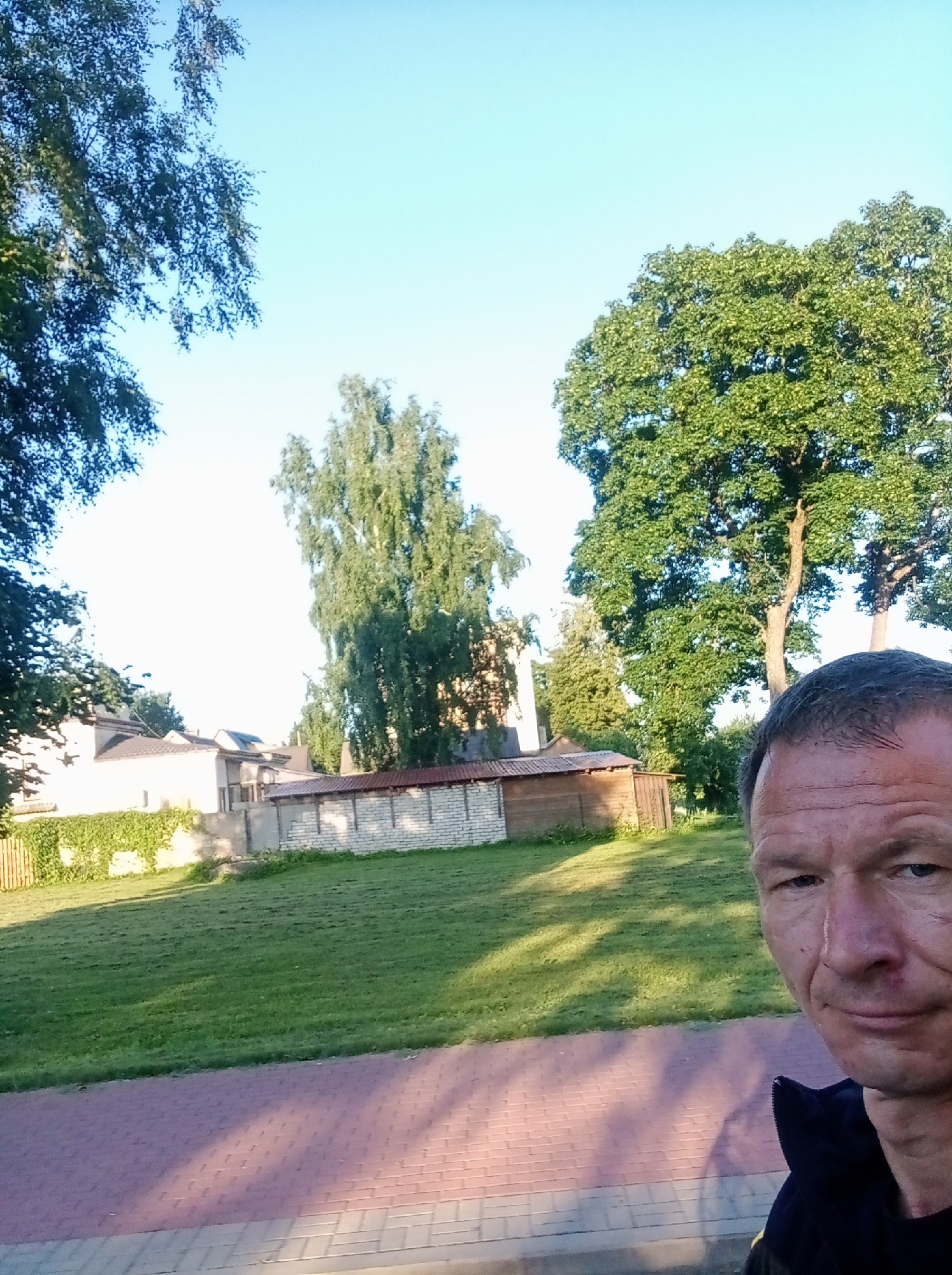 This is a selfie taken outdoors, likely on a sunny day, with a man in the foreground. The man is facing towards the camera, his head and shoulders visible. He has short, dark hair and appears to be middle-aged. He is wearing a dark jacket.
In the background, there is a grassy area, a paved walkway, and several buildings. One building is light-colored with a red-tiled roof, partially covered in green ivy. Another building appears to be a shed or garage with a wooden facade. There is also a low wall constructed from light-colored bricks. Large trees with green foliage are prominent in the midground and background, some of which are quite tall. The sky is a clear, light blue.
The scene appears to be in a residential or park-like area. The context provided suggests this location is the city of Tapa, Estonia.