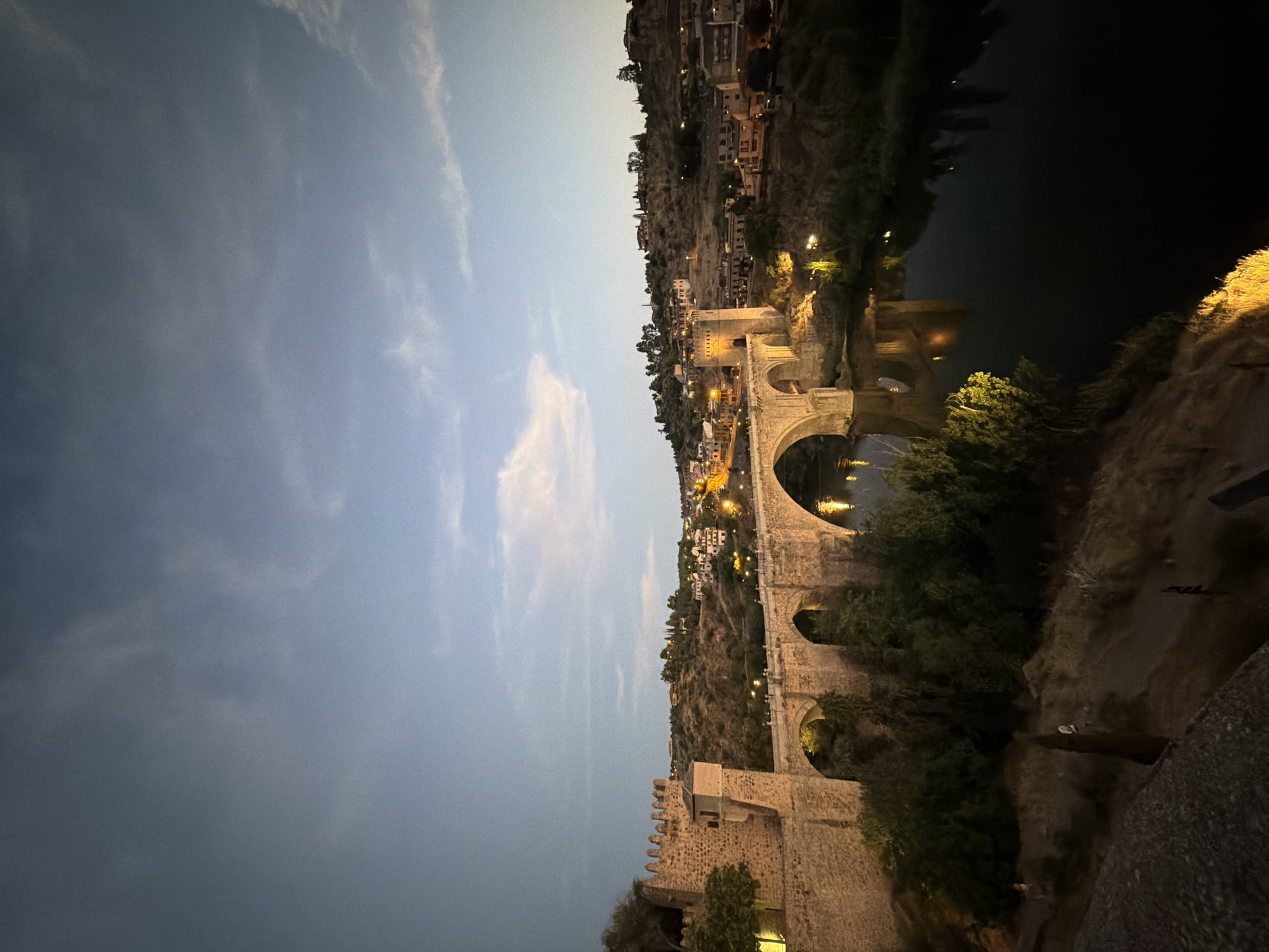 The image shows a view of the Puente de San Martín, a medieval stone bridge crossing the Tagus River in Toledo, Spain, at dusk. The bridge, constructed in the 14th century, features multiple arches and is illuminated by artificial lights, casting reflections on the dark water below. To the left of the bridge, a fortified tower with crenellated battlements stands, part of the city's ancient walls. Beyond the bridge, the hilly landscape of Toledo is visible, dotted with buildings and illuminated windows. The sky above is a gradient of twilight blue and grey, with faint wisps of clouds. The foreground consists of a textured, rocky slope with sparse vegetation. The scene is tranquil and captures the historic ambiance of the city.
