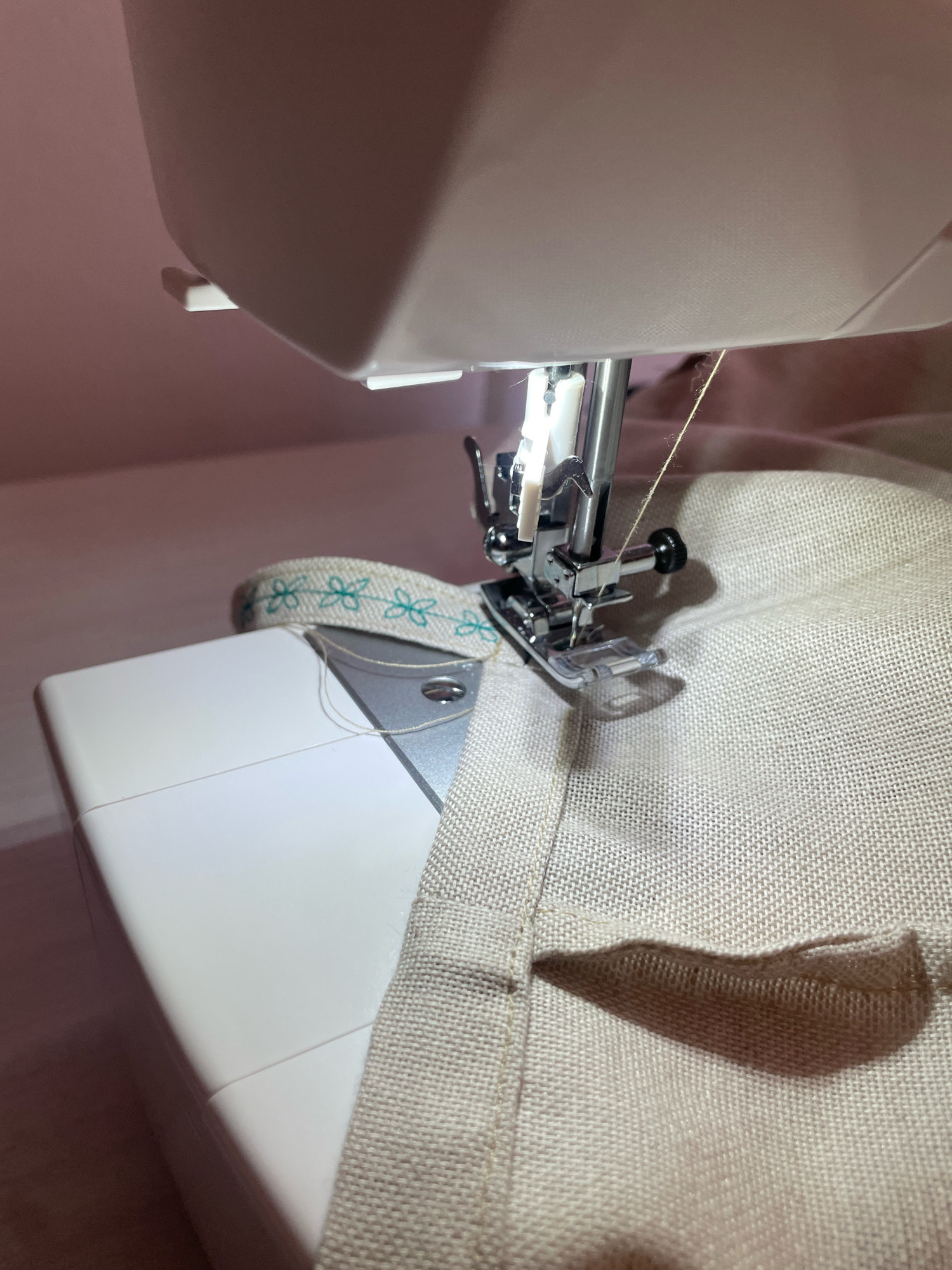 A close-up shot focuses on a sewing machine in operation. The needle, attached to the presser foot, is actively stitching beige fabric. A spool of thread is visible, with a strand of thread running from it to the needle. To the left, a white part of the sewing machine, likely the base or arm, is visible. A decorative ribbon with a floral pattern in green and white is draped over the fabric near the sewing machine. The background is a soft pink, suggesting an indoor setting. The image is static and captures a moment in the process of sewing.