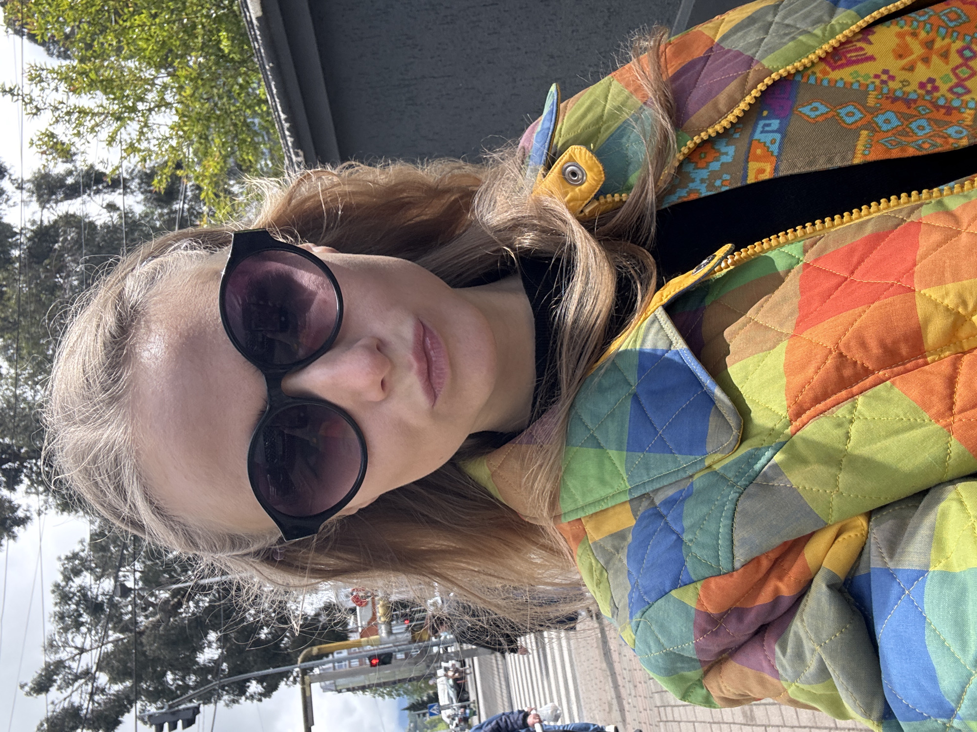 A woman wearing sunglasses and a colorful quilted jacket is standing in an urban environment. The jacket features a patchwork design with geometric shapes in various colors, and a yellow zipper. The background includes trees, a pedestrian crosswalk, and traffic lights. In the distance, pedestrians and cyclists can be observed. The location appears to be within a city, with details such as the crosswalk and traffic lights indicating urban infrastructure.