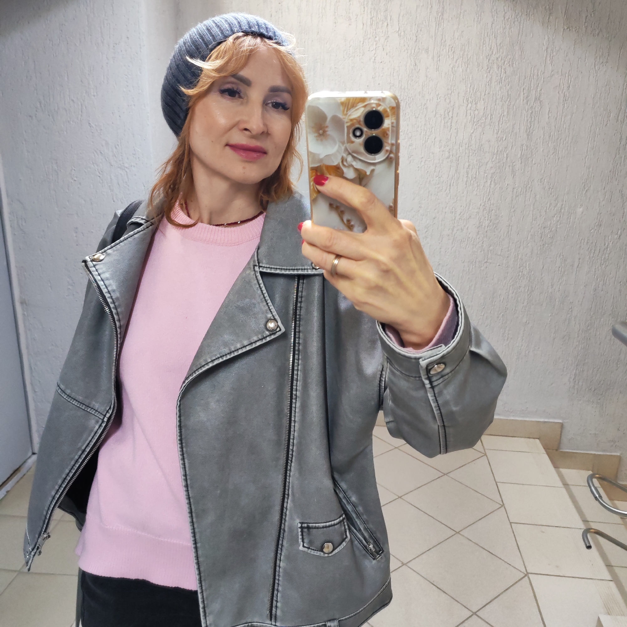 A woman takes a selfie in what appears to be an indoor setting with white walls and tiled floors. She is wearing a gray beanie, a light pink sweater, and a gray leather jacket. She is holding a cell phone with a decorative case, presumably to take the picture. The background includes architectural elements like a door and a staircase, suggesting an interior space. There is no information in the picture to confirm if the location is Chaikovsky, Russia.