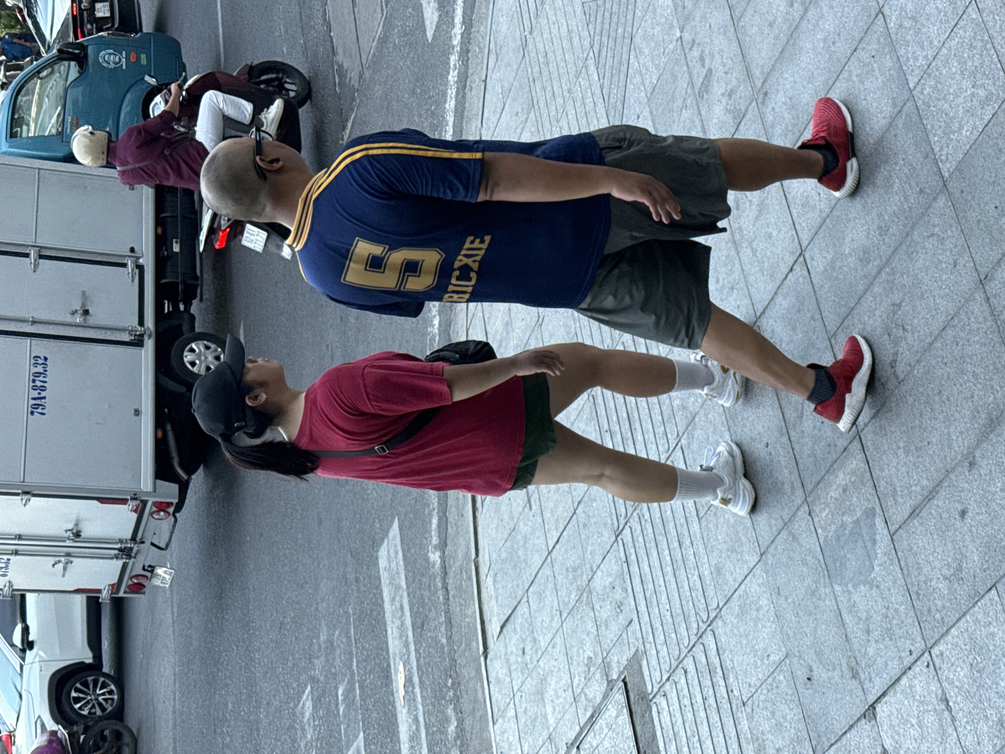 The media file shows two people walking on a sidewalk in what appears to be a city. The subjects are a man and a woman. The man is wearing a navy blue t-shirt with yellow stripes on the sleeves and "5 RIC XIE" printed in yellow on the back. He is also wearing dark shorts and red sneakers. The woman is wearing a red t-shirt, dark shorts, white socks, and white sneakers. She has a black baseball cap on her head and a black bag slung across her body.
In the background, there is a light blue truck with "79A-879.32" printed on its side. To the left of the truck is a motorcycle with a person in a maroon shirt and white pants riding it. The scene is set on a paved sidewalk with visible tile patterns. The lighting suggests it is daytime. The overall atmosphere appears casual, with the two individuals walking side-by-side. There is no discernible emotional expression from the subjects. The location is identified as Nha Trang Ward, Vietnam, which is consistent with the presence of motorbikes and the style of the truck.