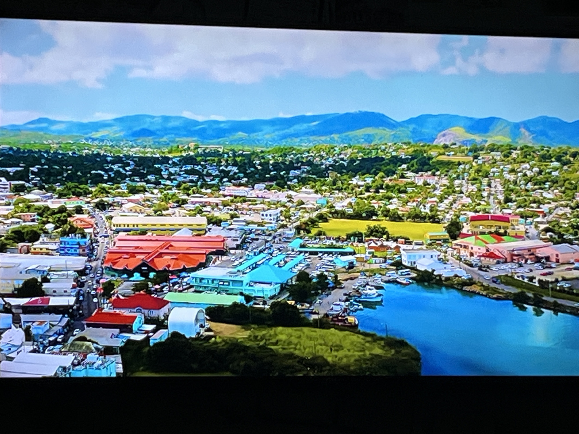 This is an aerial drone shot of a harbor in a coastal city, with mountains in the background. The city has a mix of colorful buildings, including what appear to be market stalls with bright turquoise roofs, and larger buildings with red and pink facades. There are many boats docked in the clear blue water of the harbor, as well as on land in a parking lot. The scene is vibrant and bustling with activity. The location is not Kharkiv, Ukraine.