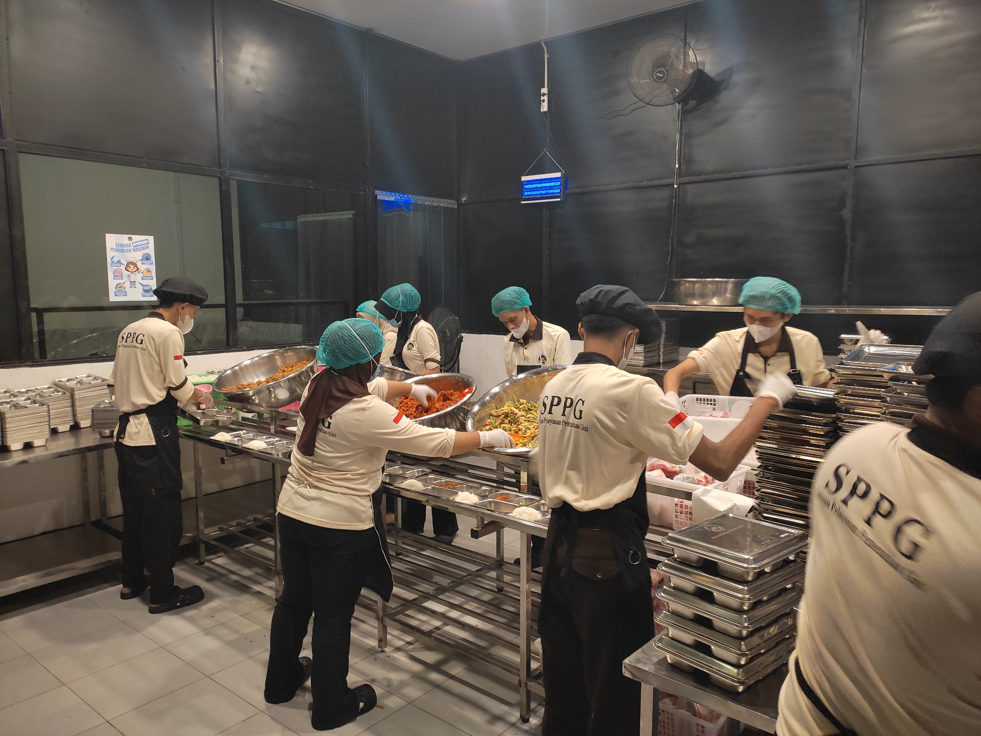 In Ponorogo, Indonesia, a group of individuals wearing hairnets and face masks are actively engaged in food preparation and serving within a commercial kitchen. Several people are seen in the foreground and midground, dressed in light-colored shirts with the "SPPG" logo and dark aprons. They are working around stainless steel counters and shelves laden with food trays and containers. Some individuals are scooping food from large metal bowls into smaller serving trays, while others appear to be organizing supplies. The environment is functional, with tiled floors and dark, paneled walls. A prominent object in the background is a wall-mounted fan. The scene suggests a busy food service operation, potentially a catering or institutional kitchen.