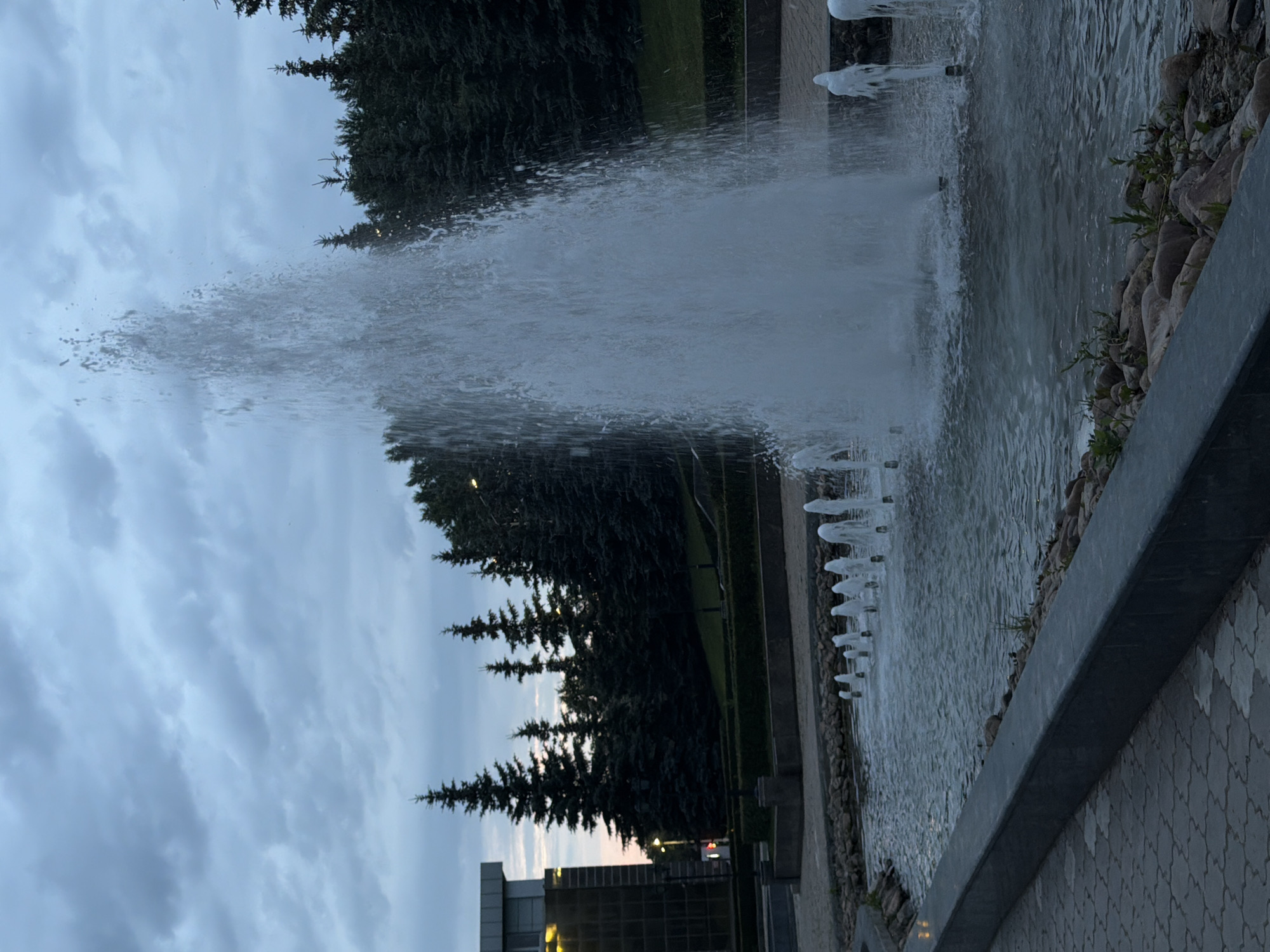 A fountain with a tall central jet and multiple smaller jets is the prominent feature in a park-like setting. The water is illuminated by a warm light source not visible in the frame, creating a shimmering effect. The fountain is surrounded by rocks and a low-lying landscaped area with manicured bushes. Beyond the fountain, a line of dark evergreen trees forms a backdrop. To the left, a modern building with large glass windows is visible. The sky above is overcast with textured clouds, suggesting dusk or a stormy day. The ground in the foreground is paved with interlocking stones.
