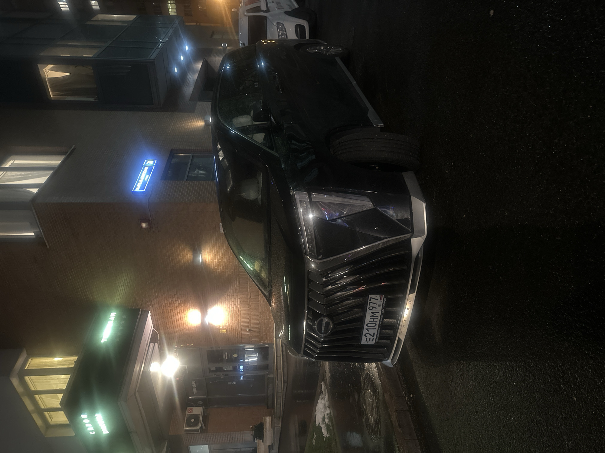 The image displays a night scene on a wet street in Khimki, Russia, featuring a prominent black SUV parked alongside a building. The ground is dark and reflective, indicating recent precipitation or wet conditions, with visible patches of snow or ice near the curb.
The main subject is a black SUV with a distinctive grille, featuring the Russian license plate "E210HM977 RUS". Below the grille, the word "MASTER" is visible on the bumper. Small text on the license plate frame hints at "GAC MOTOR" and "АВТОЦЕНТР" (Autocenter). A white van is partially visible behind the black SUV.
The building on the left has a brick facade with modern glass window sections. It is illuminated by several warm-toned streetlights. A bright blue street sign is affixed to the building, clearly displaying "УЛИЦА КАРЛА МАРКСА 8А" (KARL MARX STREET 8A), providing a specific location cue. Lower down on the building, a light-green illuminated sign is visible, with text like "CAPONE" and possibly "ТРЁХГОРКА" (Trekhgorka) or "TRE GLOW," suggesting a commercial establishment. An outdoor air conditioning unit is mounted on the building's exterior.
The scene depicts a quiet urban environment after dark, with no people visible. The presence of wet surfaces and snow suggests cold weather.