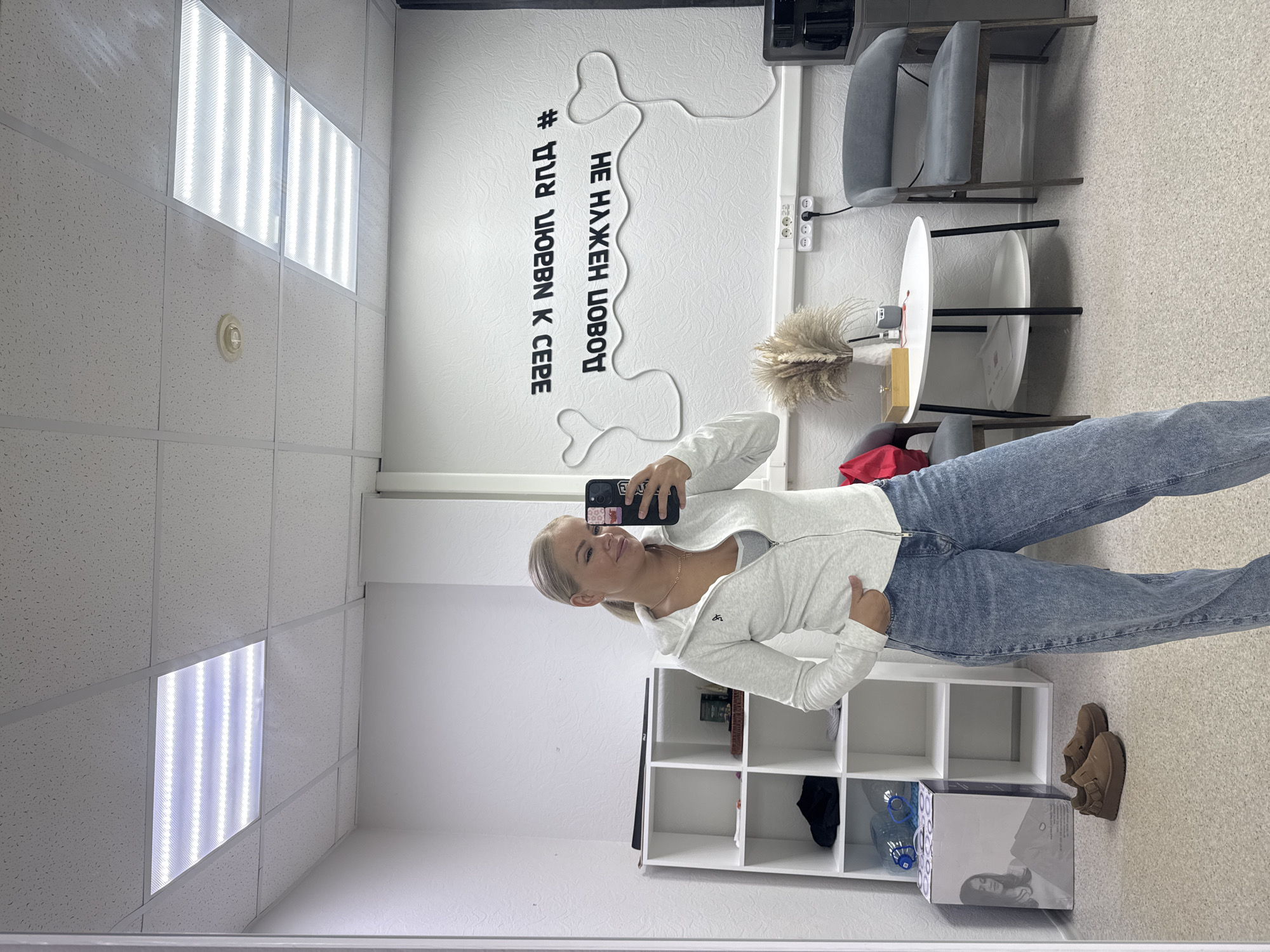A person stands in a room taking a selfie. The room has a drop ceiling with fluorescent lights. There is a white shelving unit, shoes, and a box on the floor. On the wall is the text "# ДЛЯ ЛЮБВИ К СЕБЕ НЕ НАЖЕН ДОВОД." There is a table with a vase on it and two chairs. The location is an interior space in the city of Unknown, Unknown.