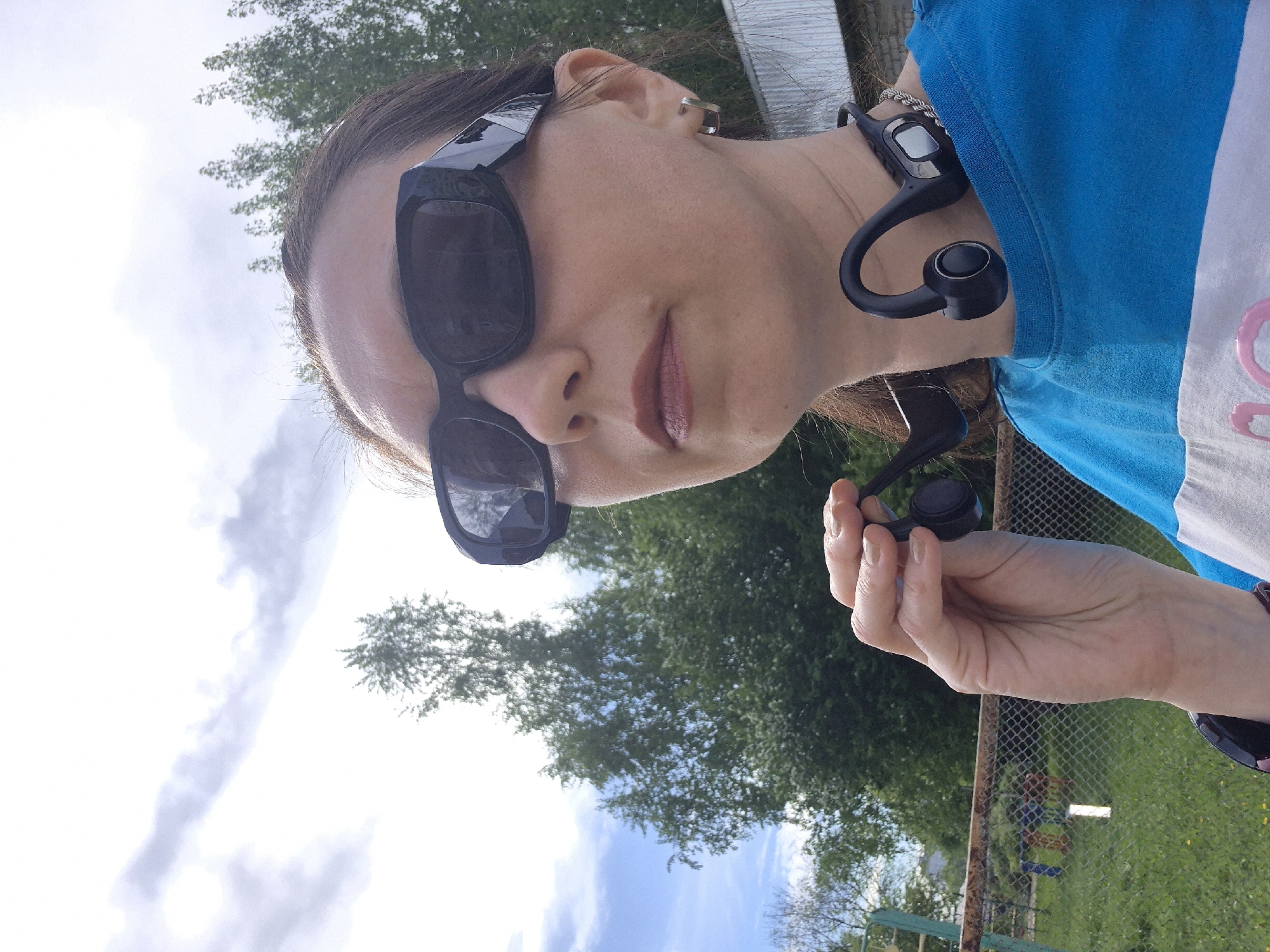 A woman wearing sunglasses and a blue shirt is outdoors. She is holding a black wireless headset in her hand, while a similar device is around her neck. The setting appears to be a yard or park with trees and a fence visible in the background. The sky is partially cloudy.