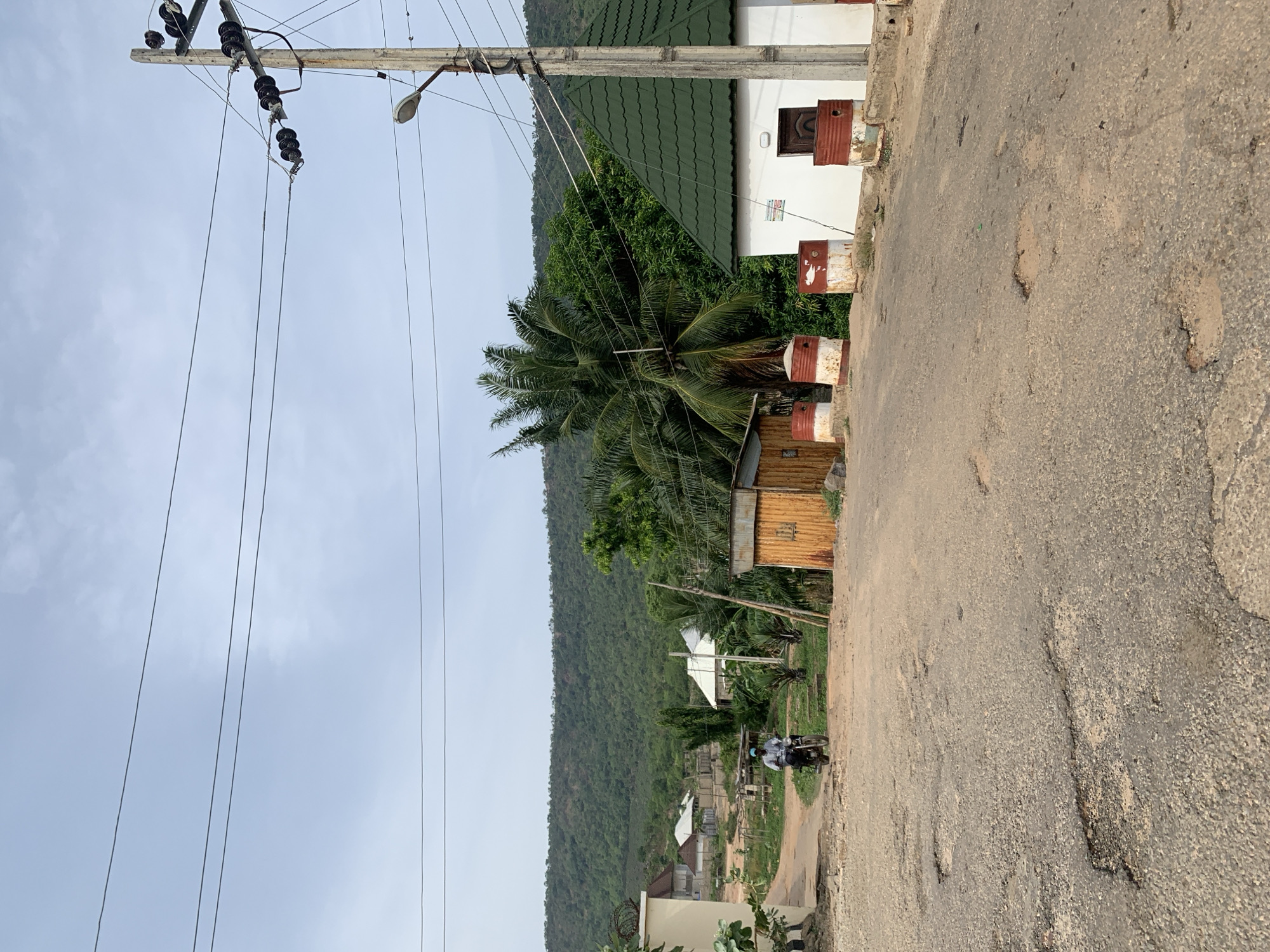 A road in Lokoja, Nigeria, shows a worn asphalt surface with numerous potholes. An overhead power line stretches across the upper-left quadrant. A street lamp is attached to a utility pole. A white building with a green roof and red-and-white painted metal drums serving as makeshift bollards is on the right. A palm tree and other vegetation are nearby. In the distance, a green, densely vegetated hill is visible. A motorcyclist is on the road further away.