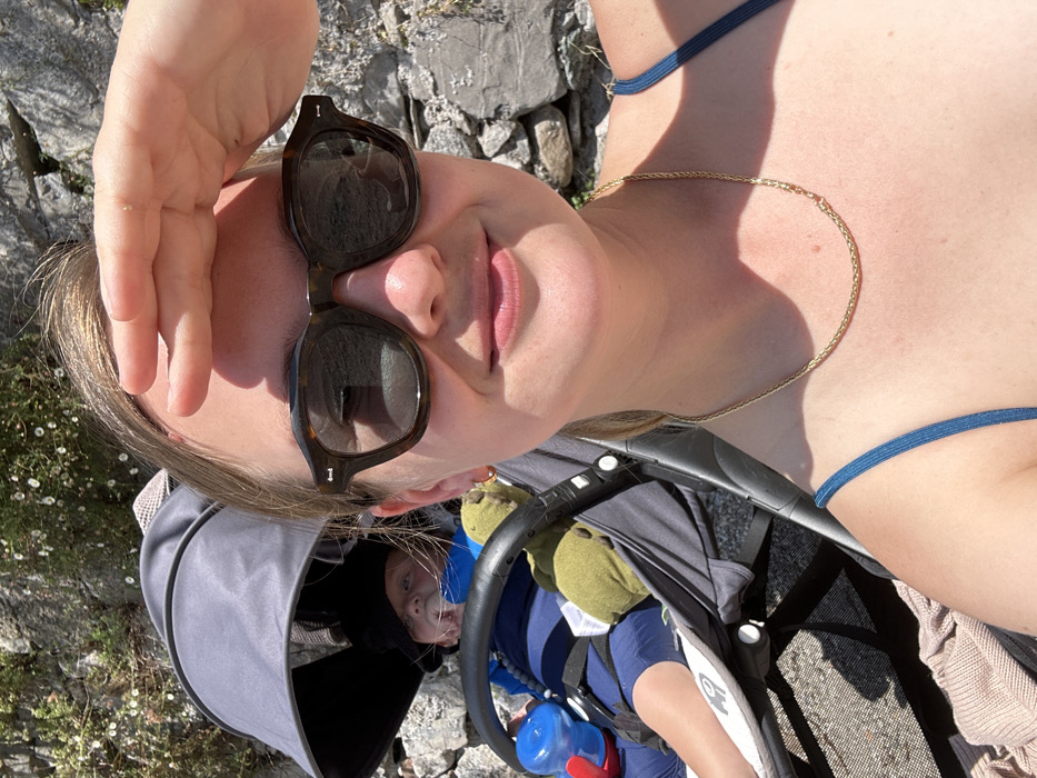 A selfie-style photograph captures an adult female and an infant in a stroller, situated outdoors in Abbadia Lariana, Italy. The female, positioned in the foreground, wears dark, tortoise-shell sunglasses and a gold-colored chain necklace over a dark blue-toned garment strap. Her right hand is raised to shield her eyes from the bright sunlight. Her expression displays a subtle smile. To her left and slightly behind her, an infant is seated in a gray stroller equipped with a dark canopy. The infant wears a dark head covering, and a blue sippy cup with a red spout is visible on the stroller's left side. A green stuffed animal rests next to the infant. The background consists of a sunlit rock face with sparse vegetation, including small white flowers. The ground visible beneath the stroller is dark, partially covered by a light-colored textile.